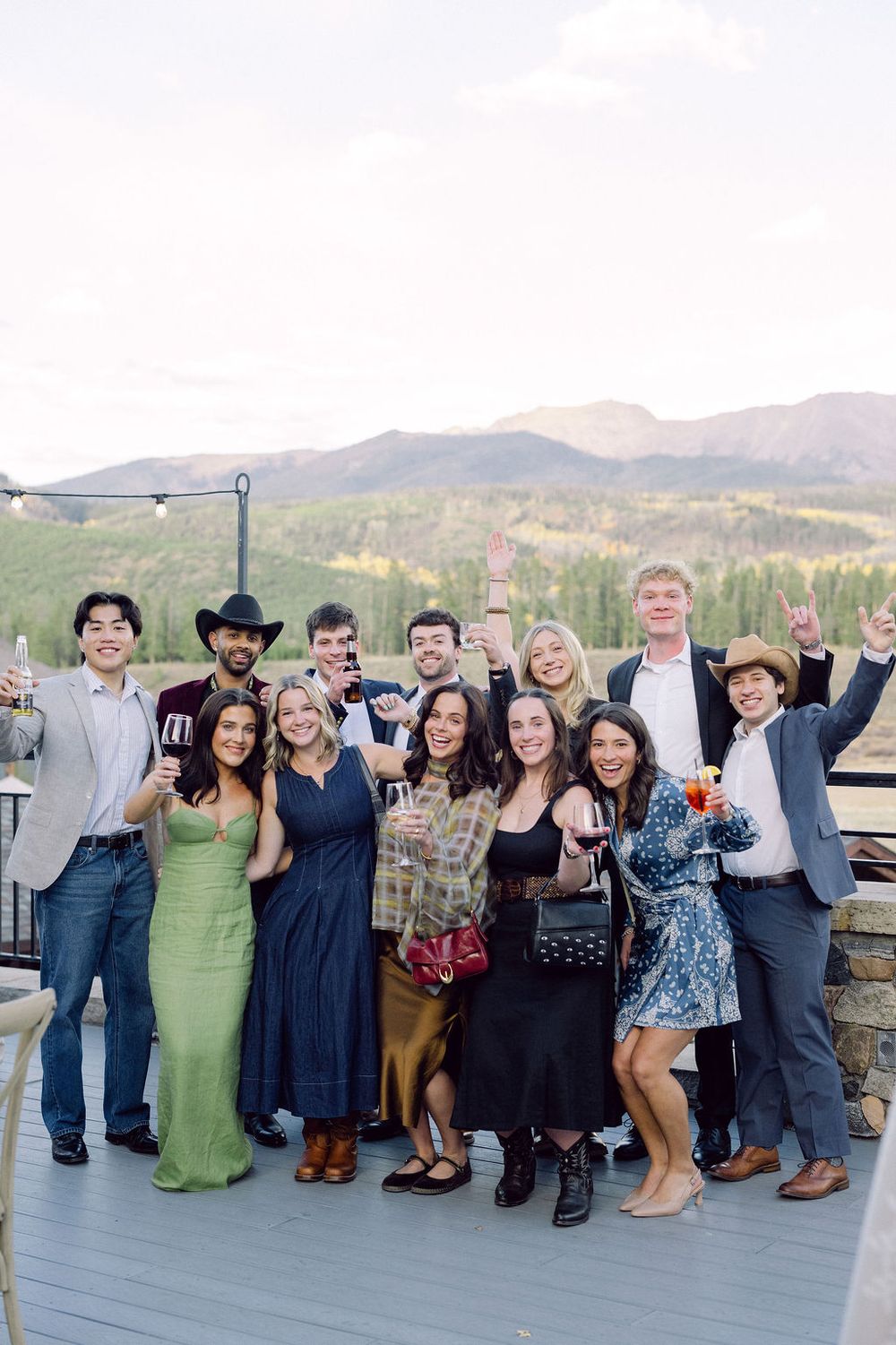 A large group of wedding guests pose together on an outdoor deck with mountain views in the background.