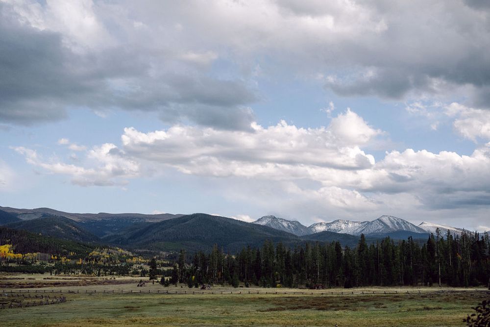 Scenic mountain landscape with rolling hills and scattered evergreen trees under dramatic cloudy skies.