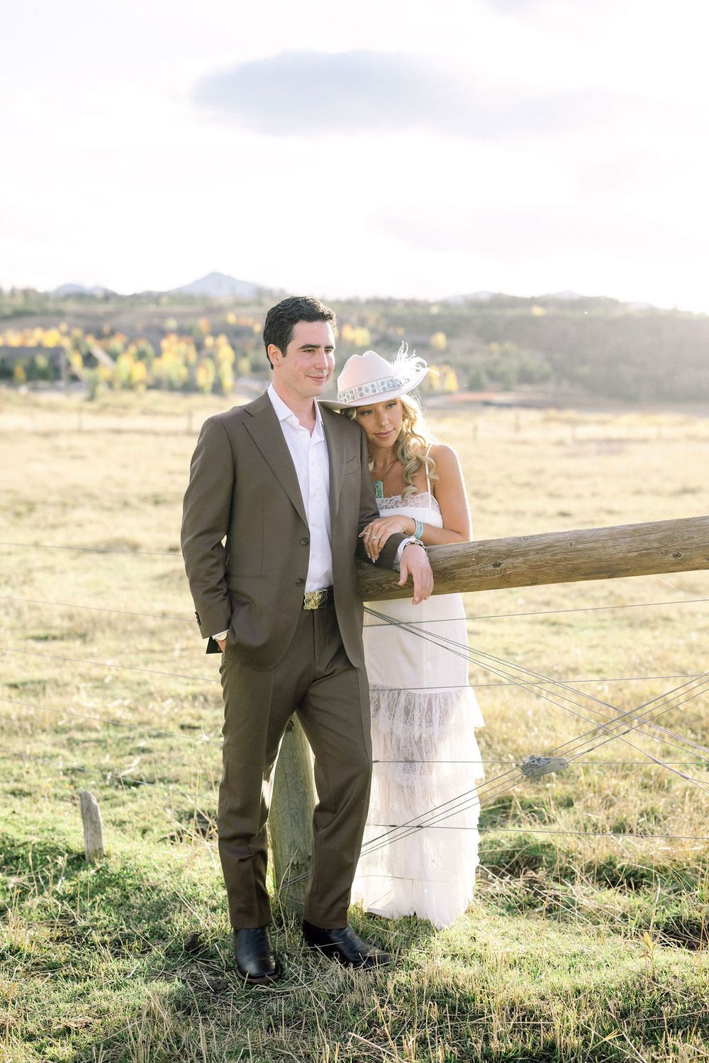 A romantic couple poses together in a sunlit meadow with mountains in the background during golden hour.