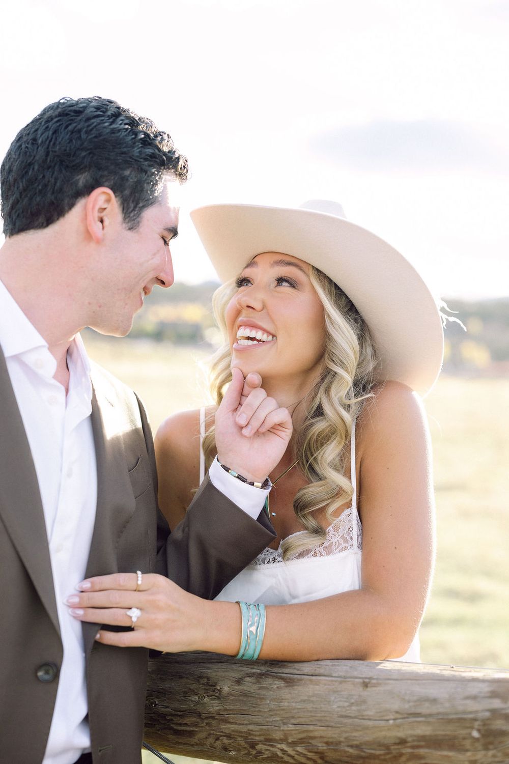 A couple shares a joyful moment together outdoors, with the bride wearing a wide-brimmed hat and western-style jewelry.