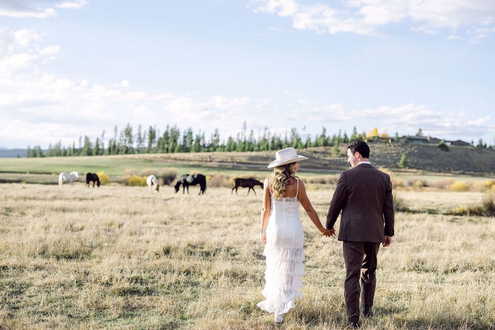 A couple in wedding attire walks through a scenic golden field during sunset in a rustic outdoor setting.