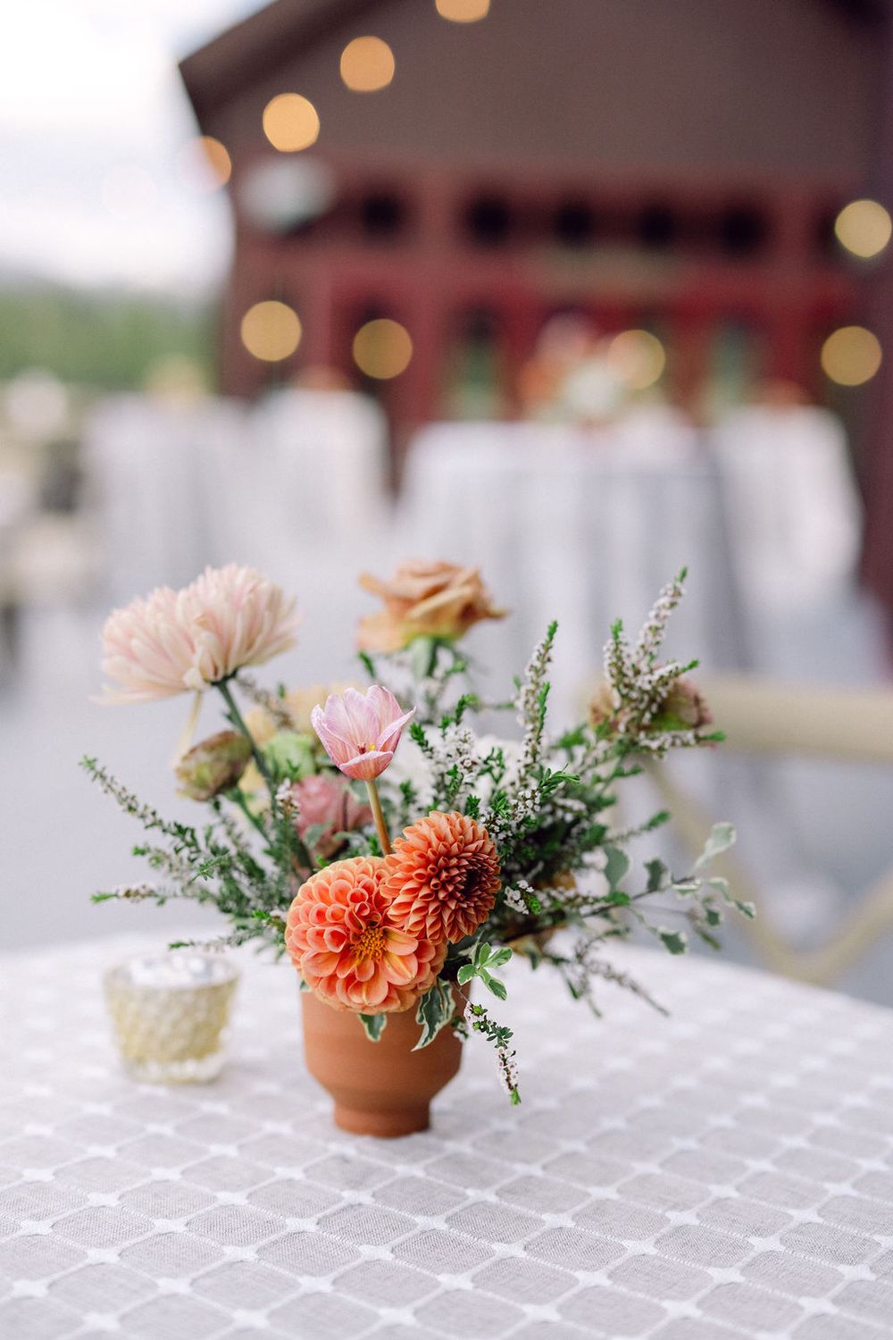 Small floral centerpiece with coral dahlias and delicate greenery arranged in a terra cotta pot on white tablecloth.