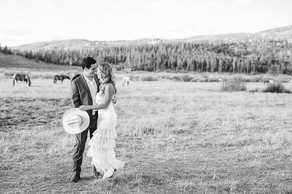 A couple embraces in a scenic outdoor field with mountains and forest in the background.