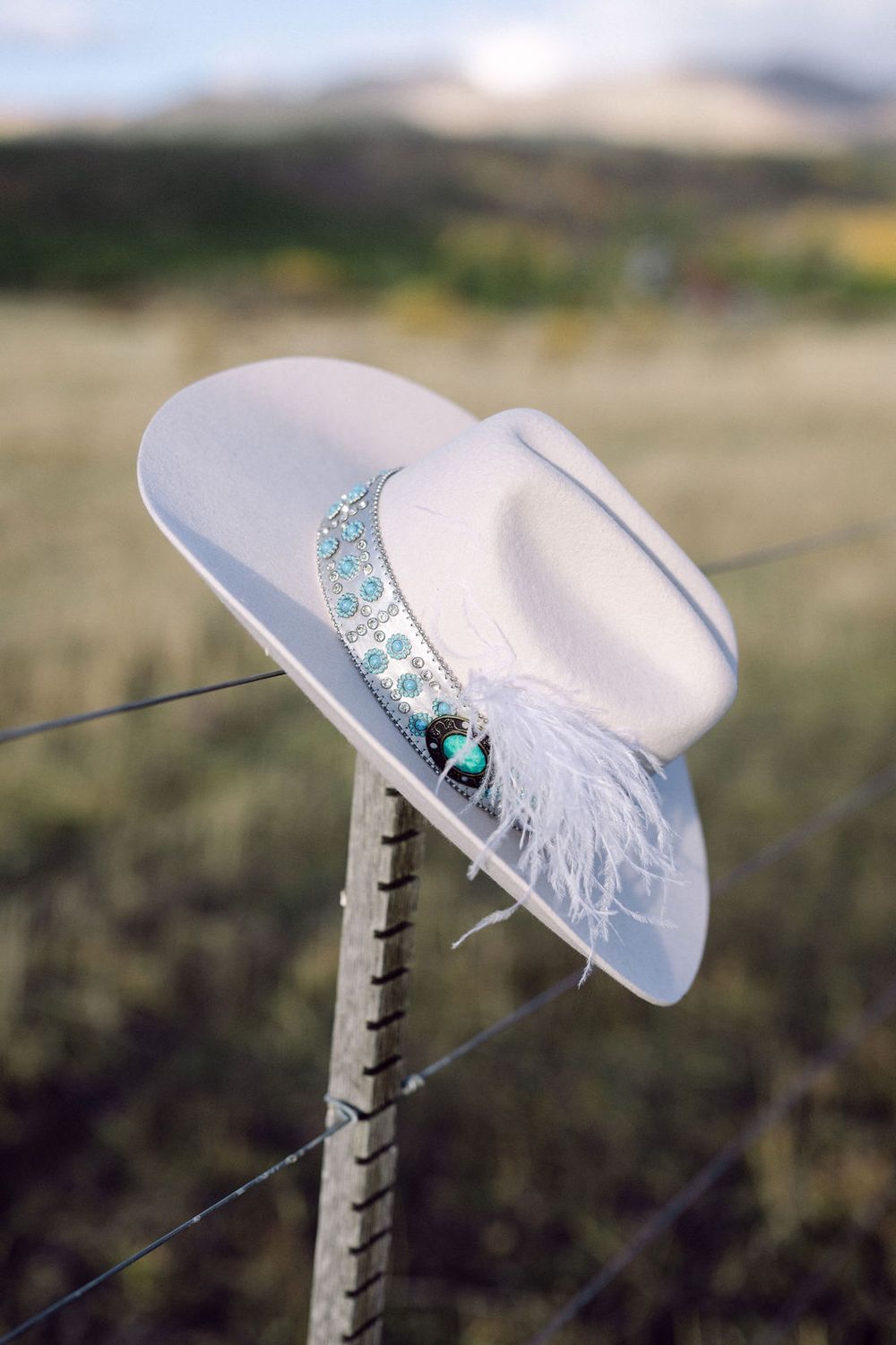 A white bridal hat adorned with sparkling blue jewels and feathers rests on a wooden post in an outdoor setting.