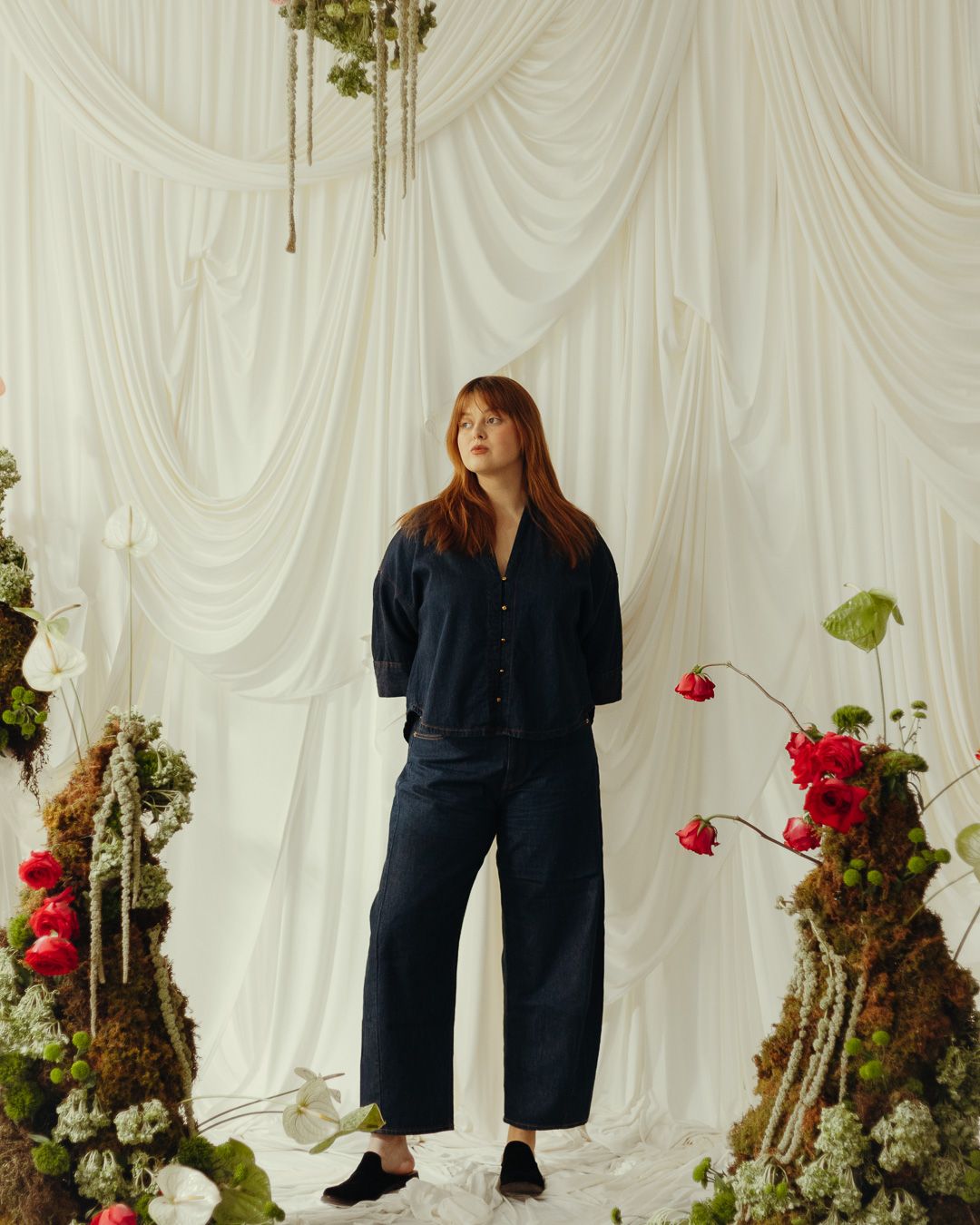 A person in a navy blue outfit poses against a white draped backdrop with red flowers.
