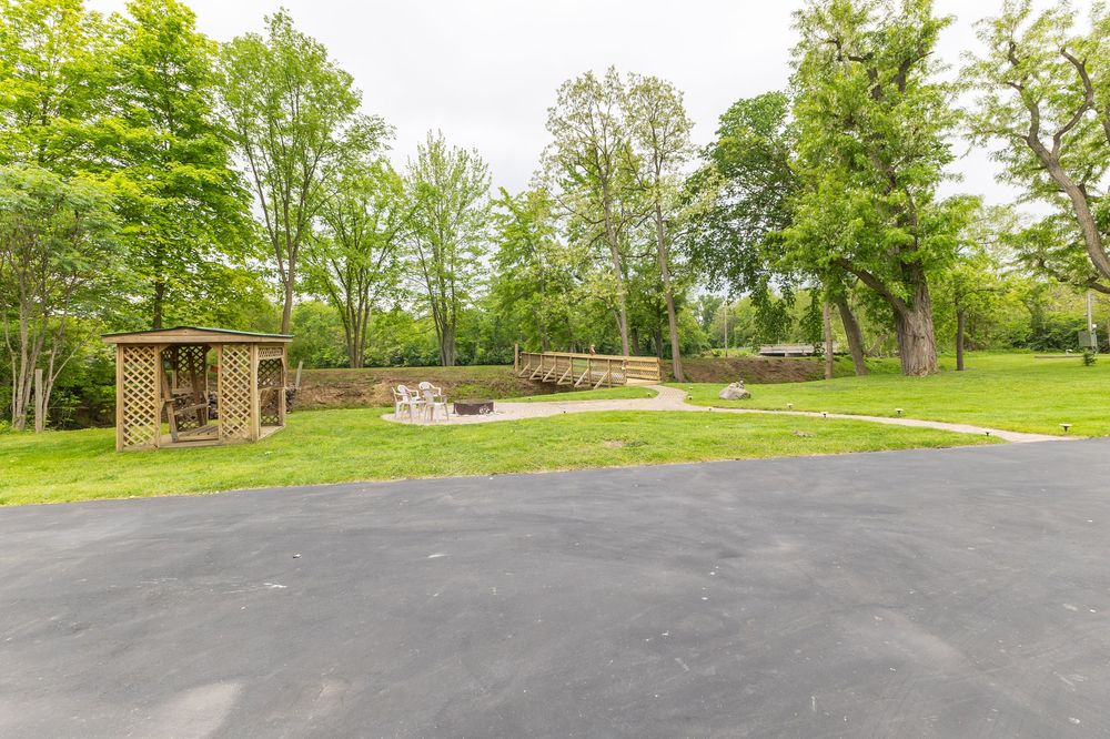 Spacious backyard with lush green lawn, mature trees, wooden pergola, and stone patio area on a sunny day.