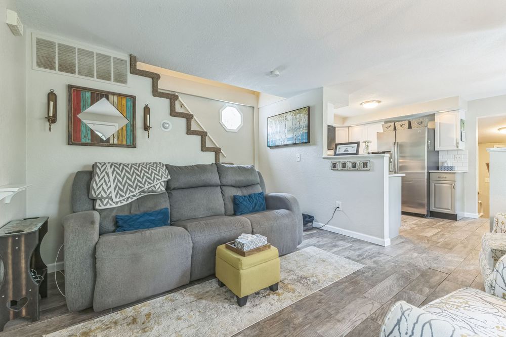 Bright living room with gray reclining sofa, light hardwood floors, staircase, and open view to modern kitchen.