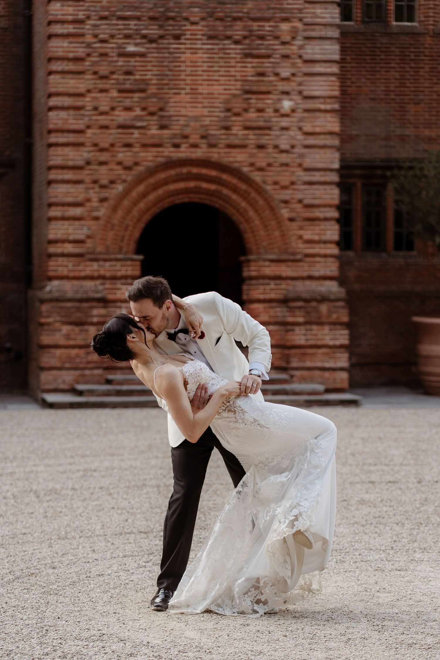 A romantic couple in wedding attire share a dramatic dip pose against a brick building backdrop.