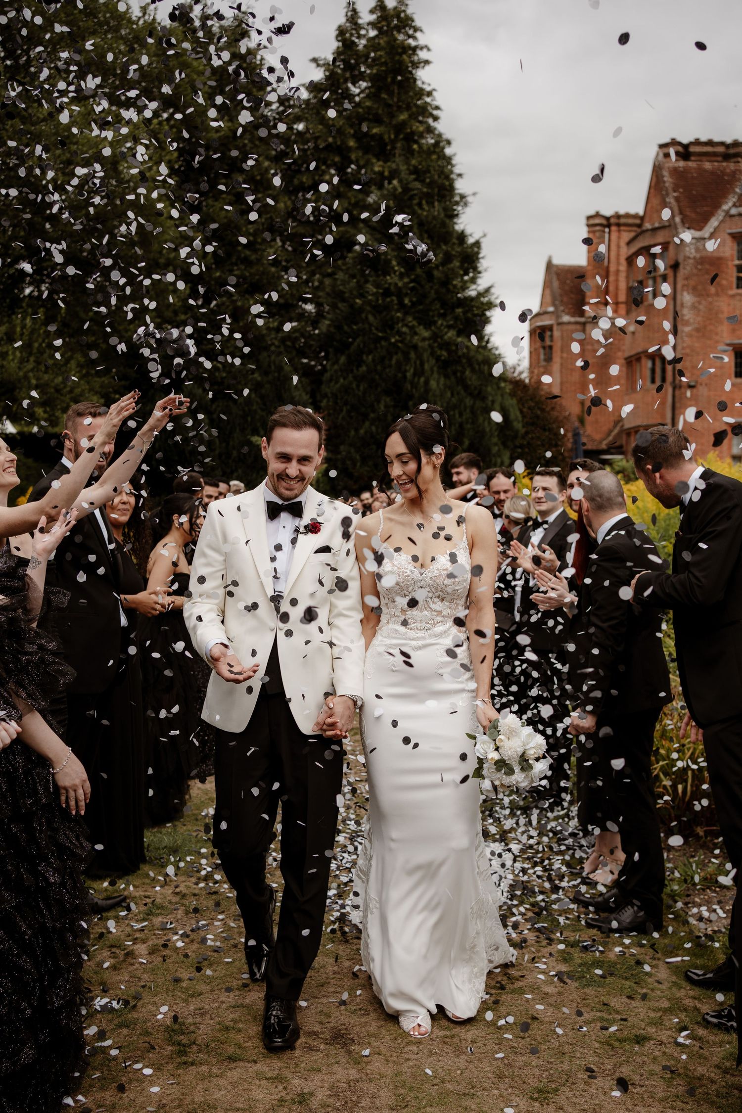 Newlyweds walk through shower of white confetti outside brick building during wedding celebration.