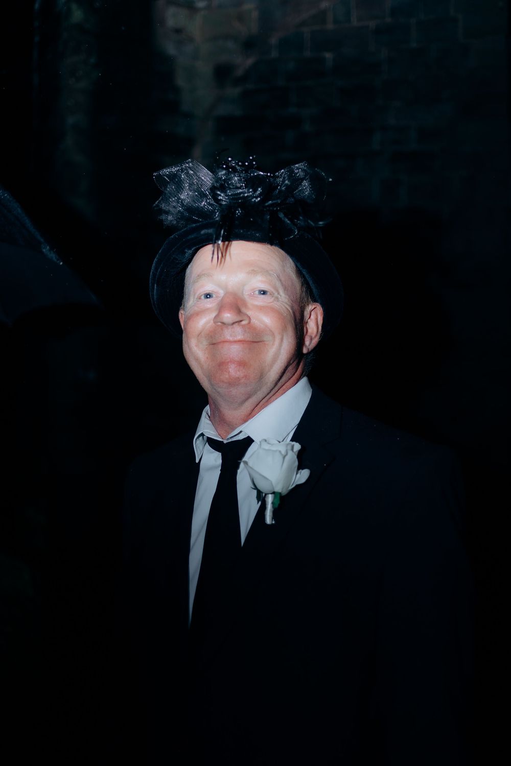 A person wearing a black hat and suit smiles warmly at the camera in dark lighting.