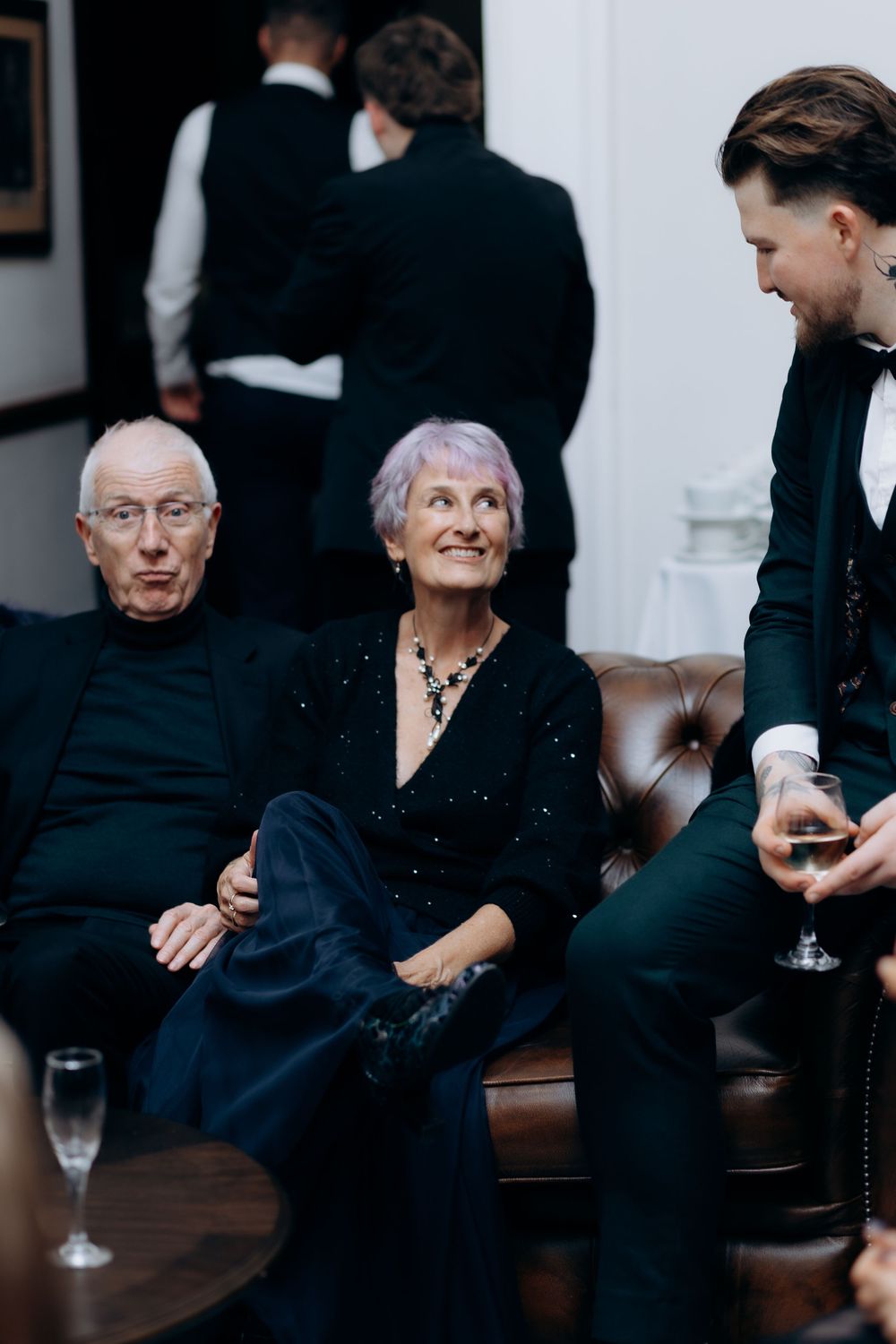 Elegant gathering shows people relaxing on a brown leather couch while being served drinks at an upscale event.