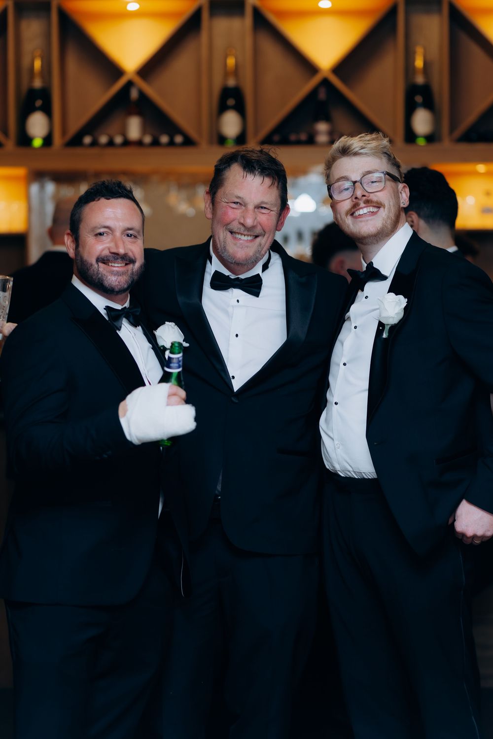 A group of men in formal black tuxedos and bow ties pose together at a wedding reception.