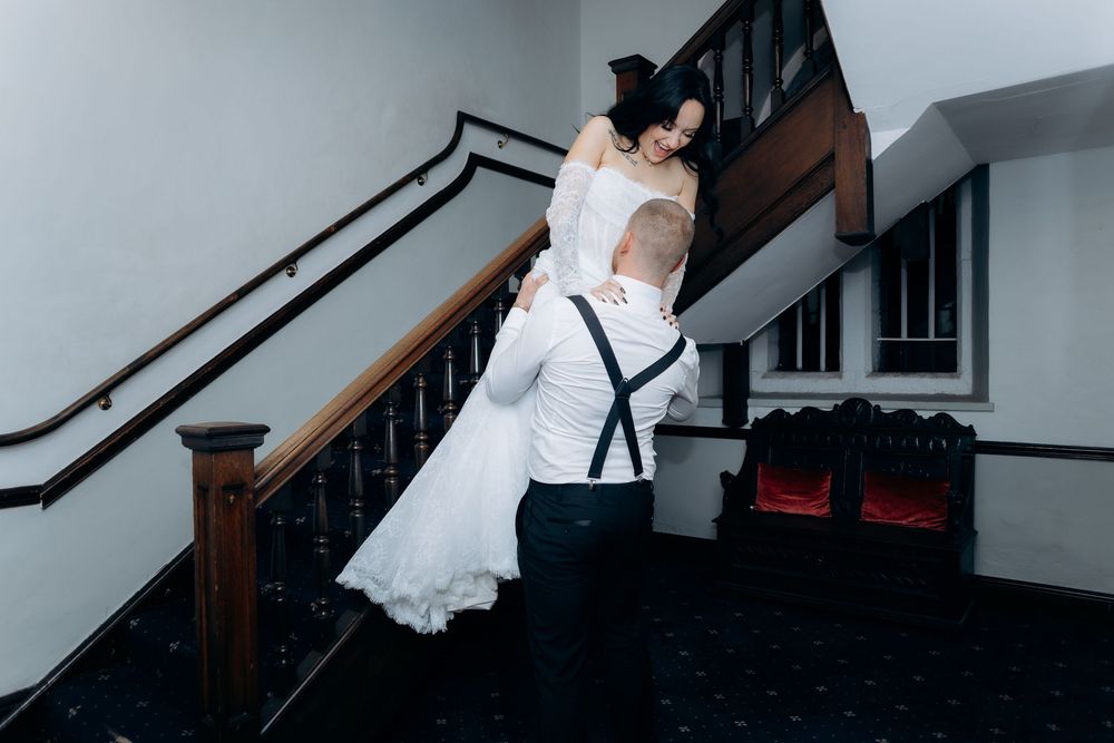 A couple in formal wedding attire embraces on a dark staircase with white walls and wooden railings.