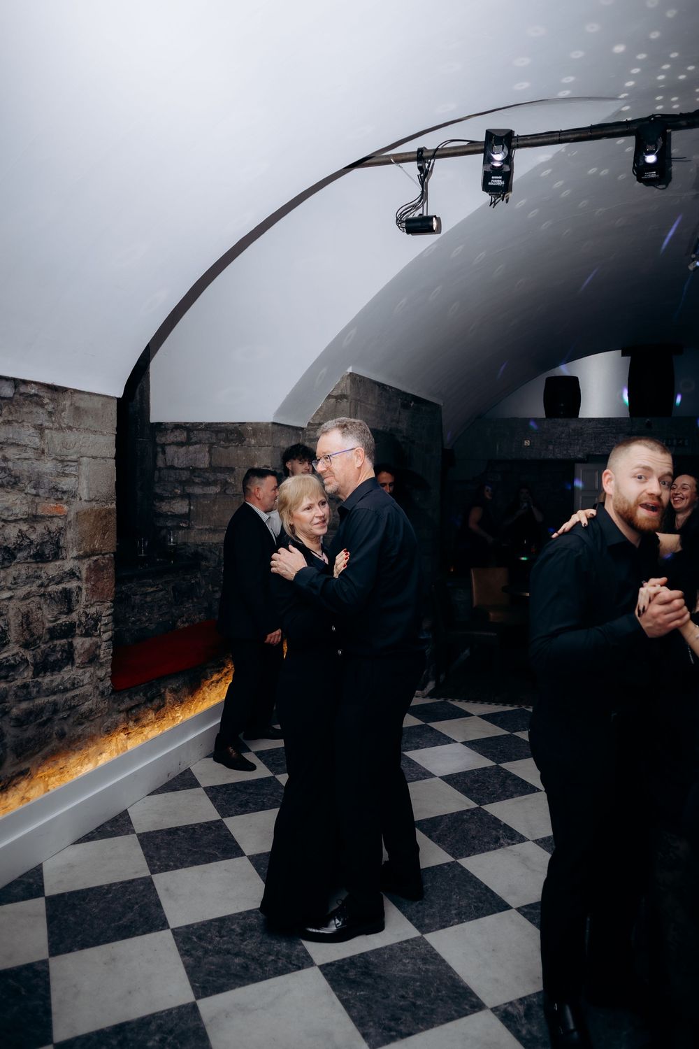 Couples dancing together in a dimly lit venue with black and white checkered floor and brick walls.