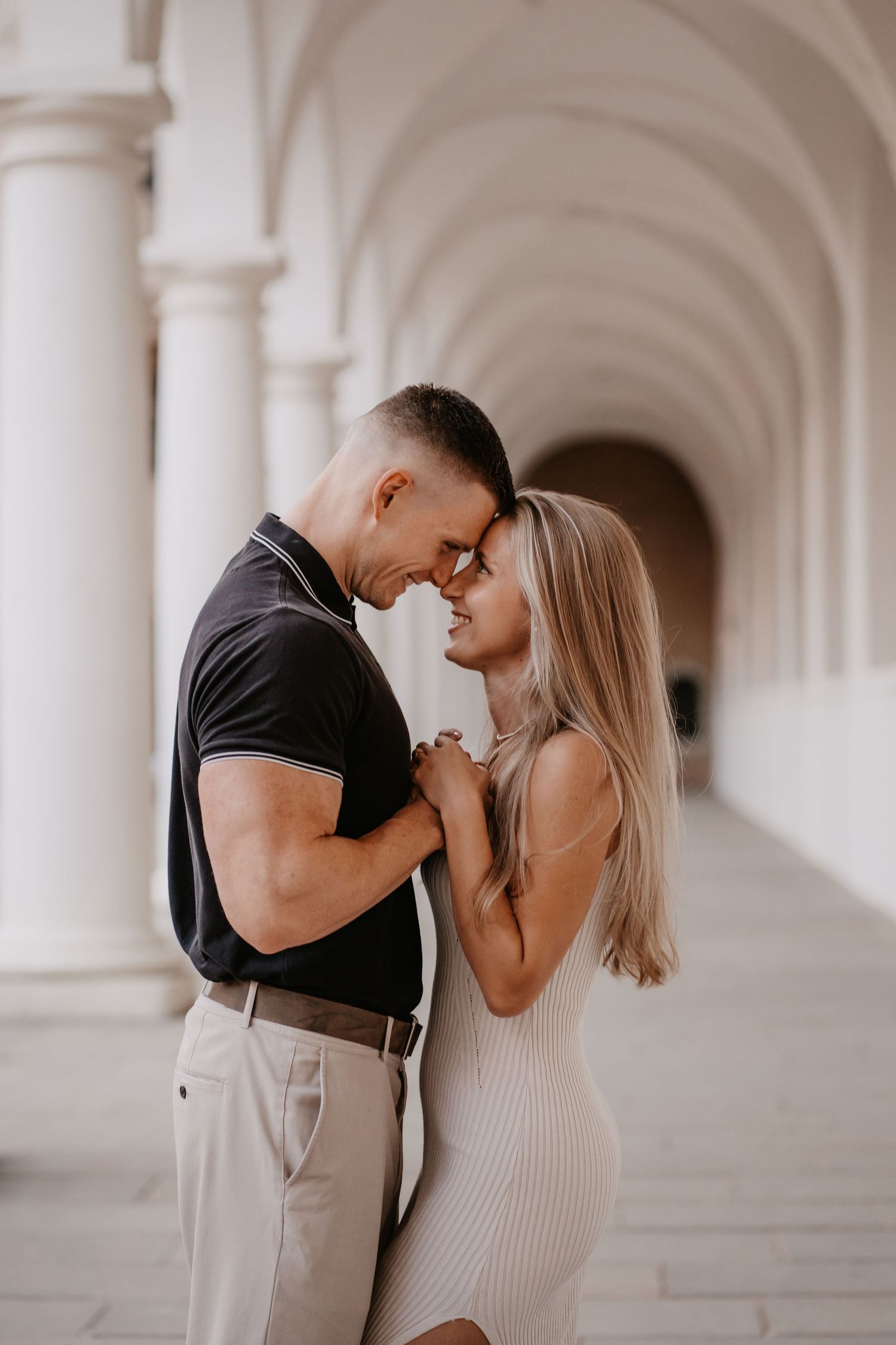 Romantic couple shares an intimate moment in an elegant white archway corridor with soft lighting.