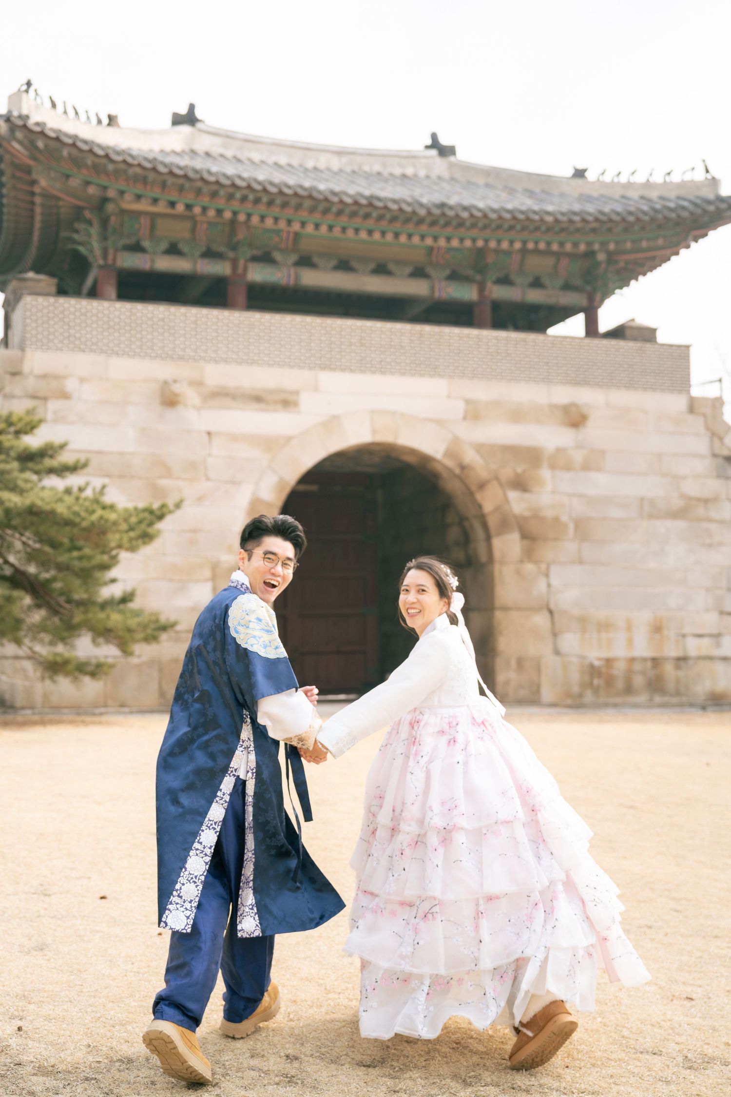 A couple in traditional Korean hanbok clothing playfully pose together at Gyeongbokgung Palace courtyard.