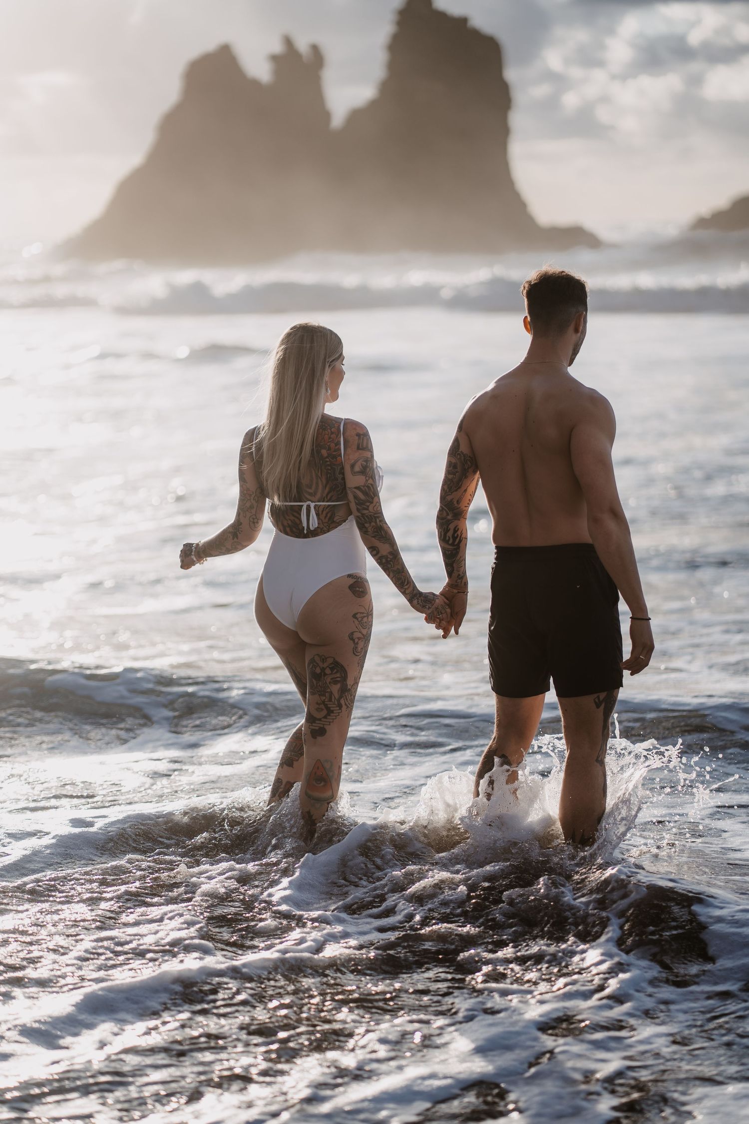 Couple wading through ocean waves at sunset with dramatic coastal rock formations in the background.