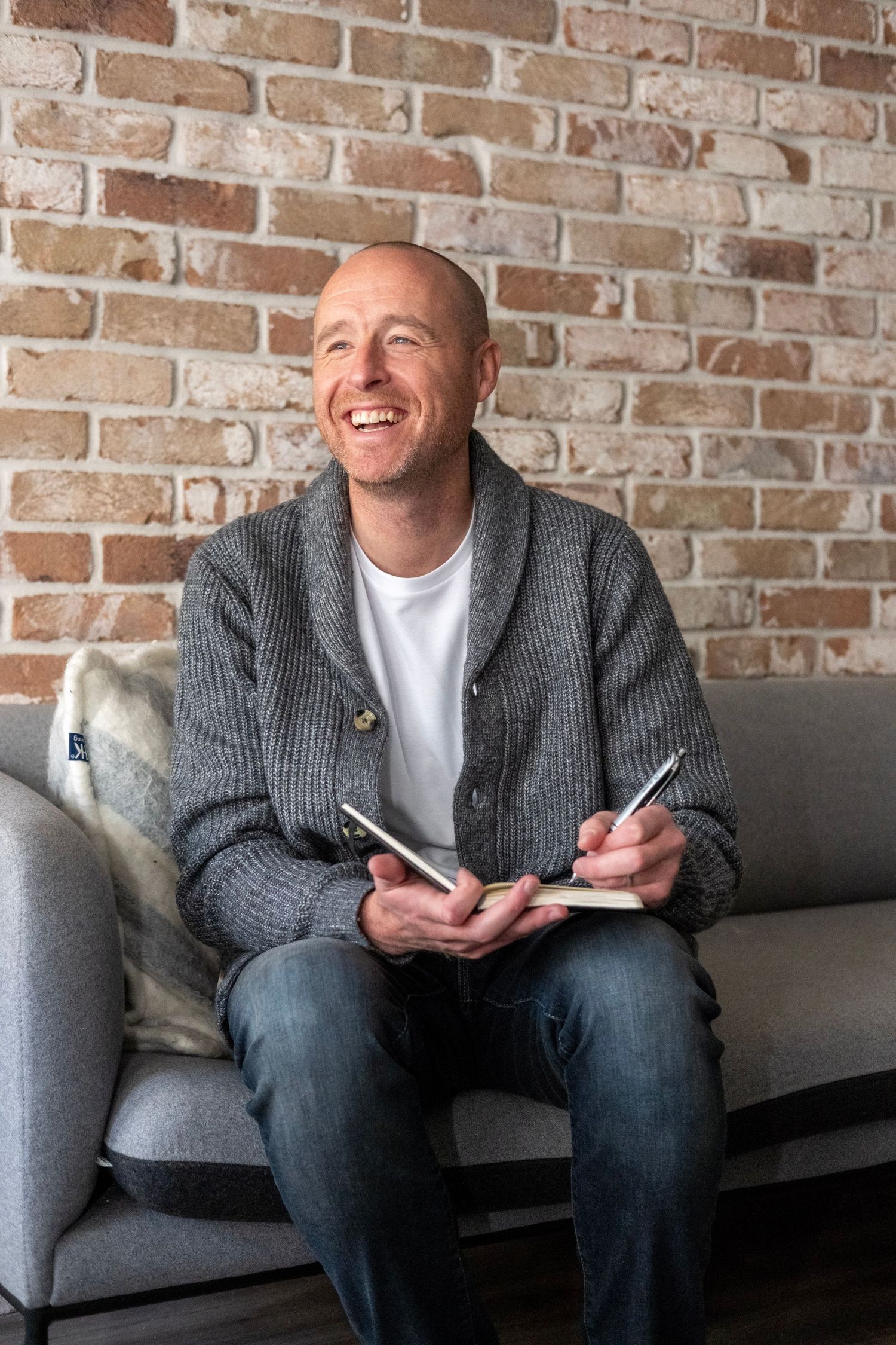 A person sitting on a gray couch against a brick wall smiles while looking at a mobile phone.