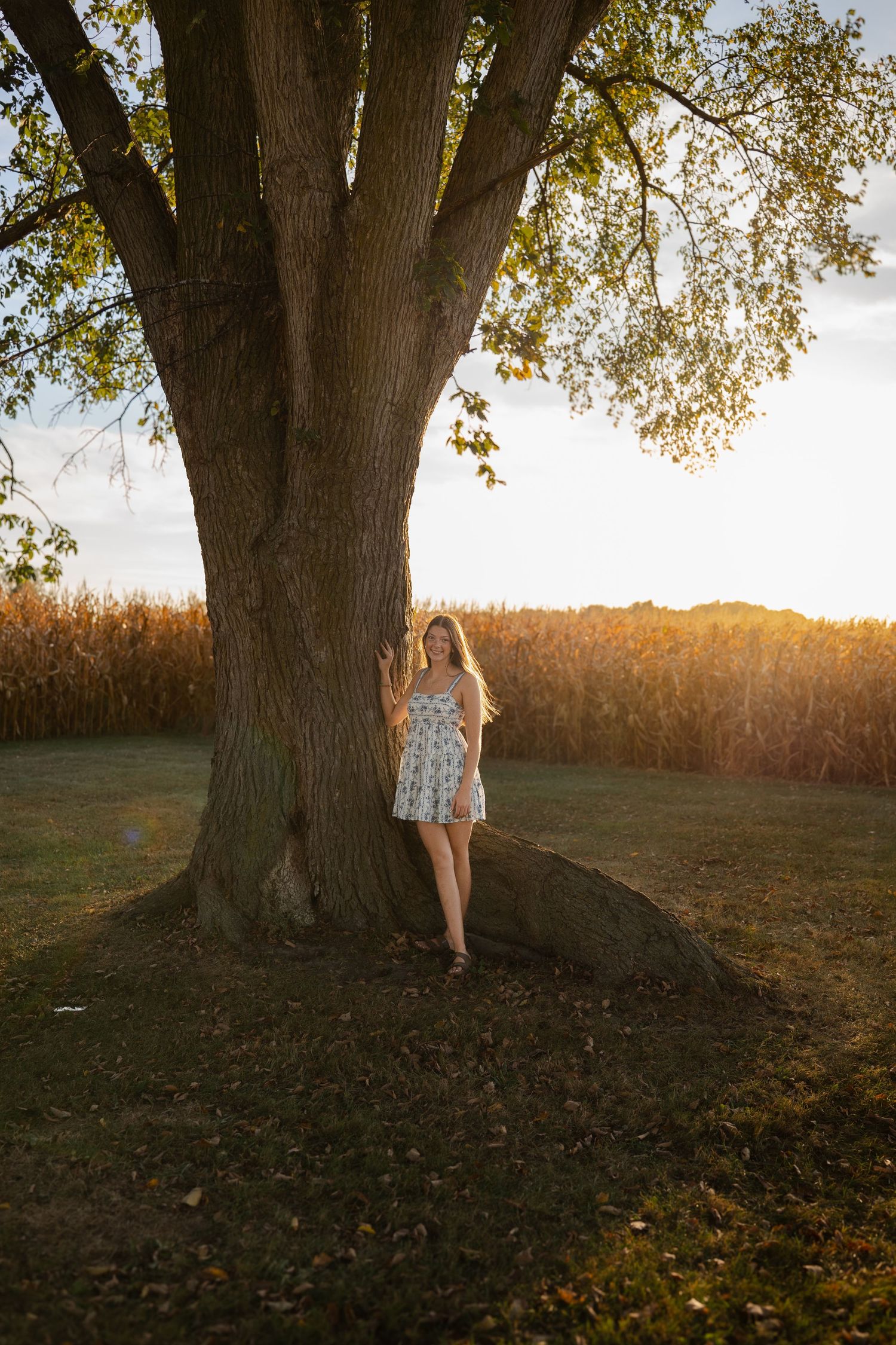 Person in floral dress poses by large tree trunk at sunset with cornfield in background.