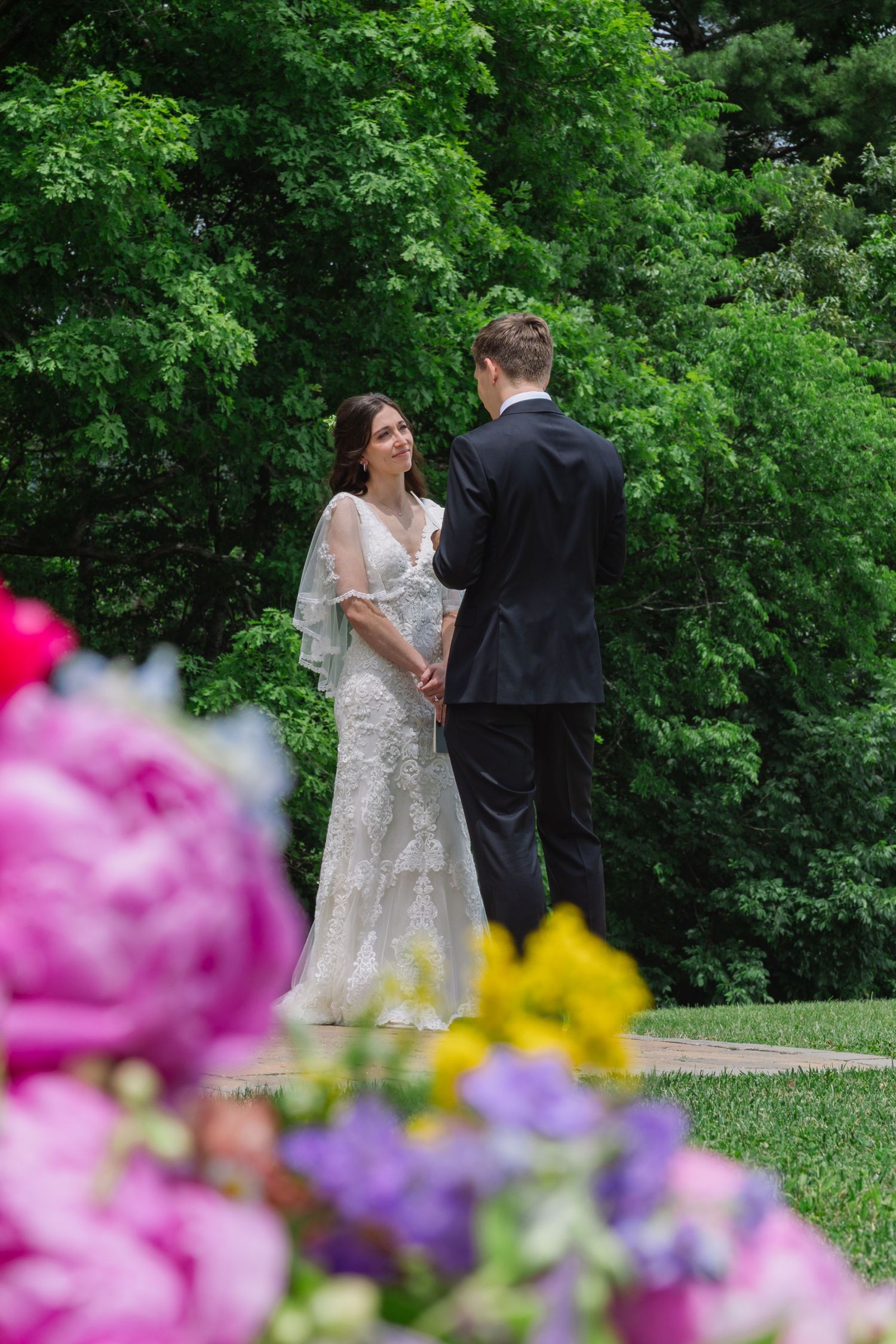 A couple in wedding attire walks along a garden path framed by colorful pink and yellow flowers in the foreground.