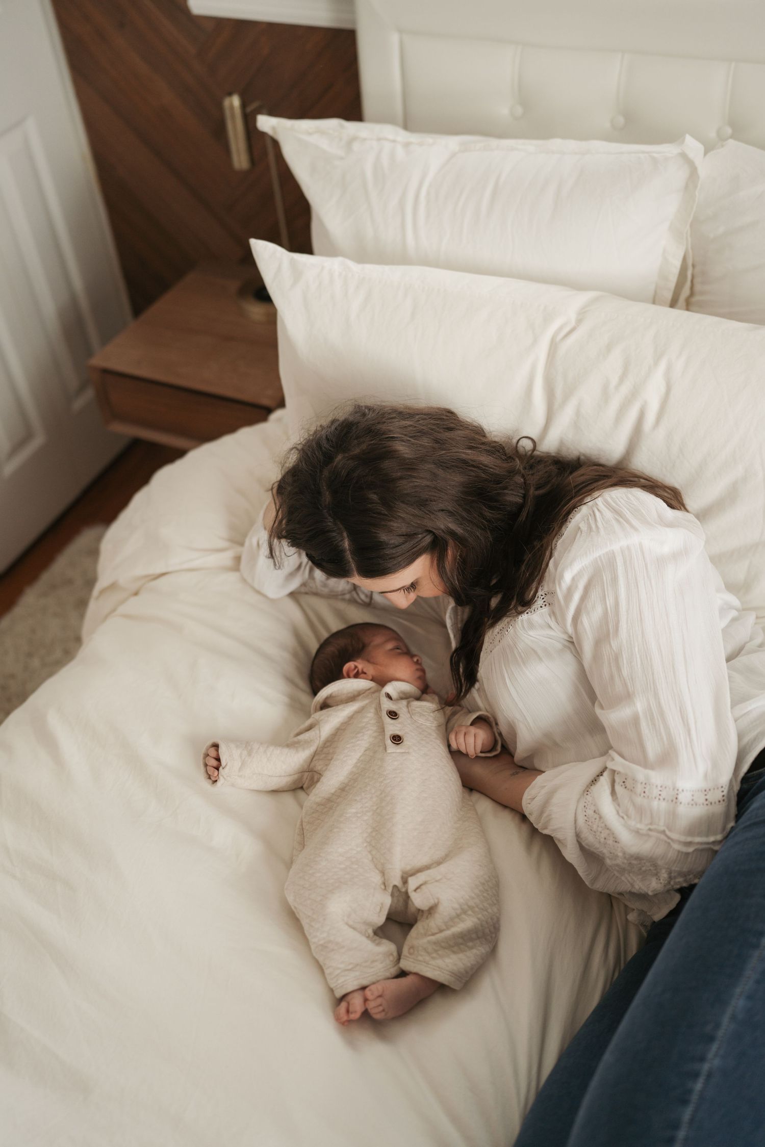Mom cuddling a newborn in white bedding while wearing a cozy sweater at in home newborn session
