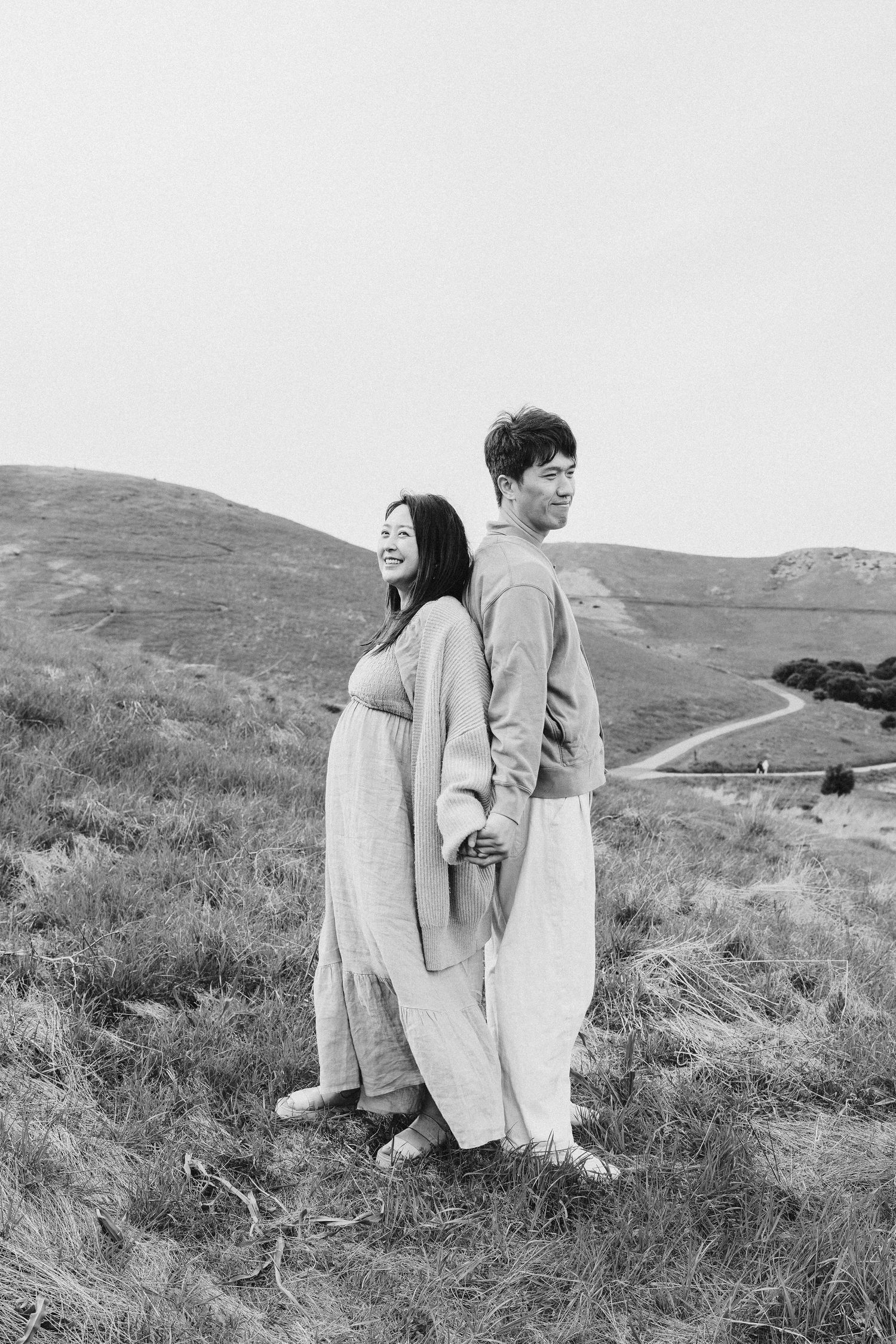 A couple in light-colored outfits stands back-to-back on a grassy hillside in black and white photography.