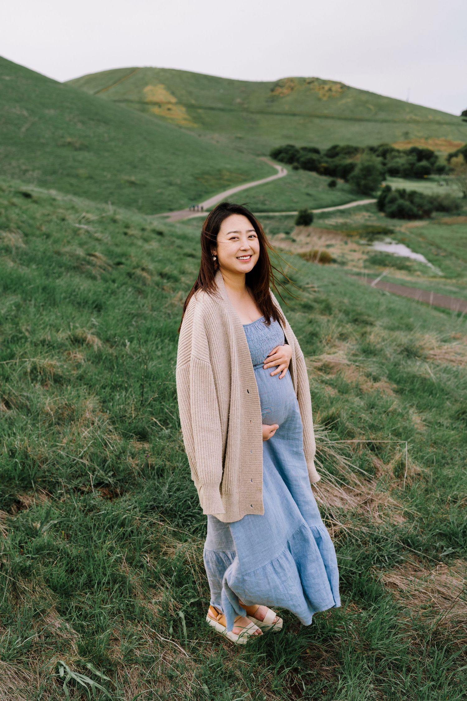 A person in a blue dress and beige cardigan sits on a grassy hillside during golden hour.
