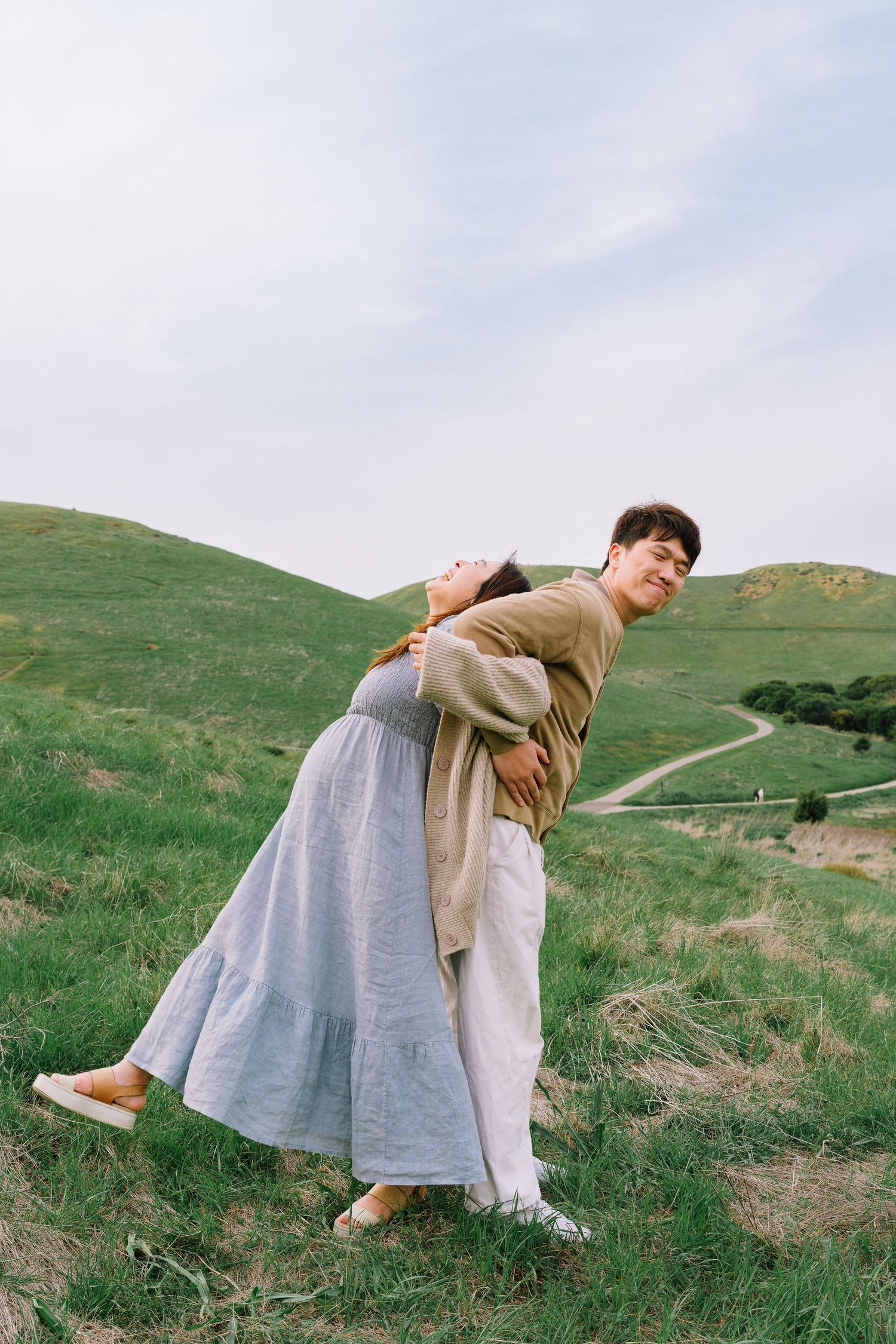 A couple dances playfully on a green hillside trail during a sunny afternoon.