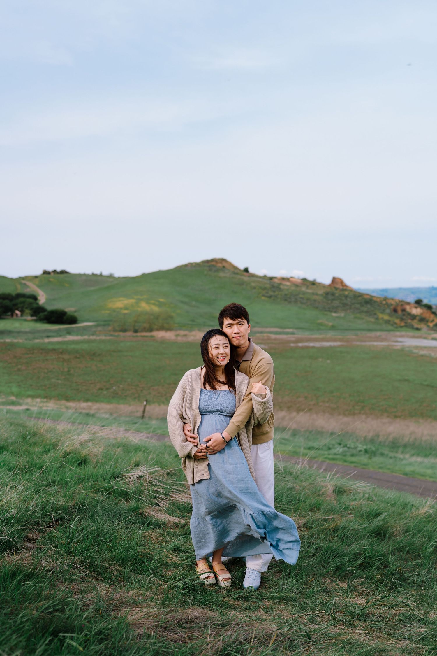Two people share an intimate moment while sitting together on grassy hills under a blue sky.