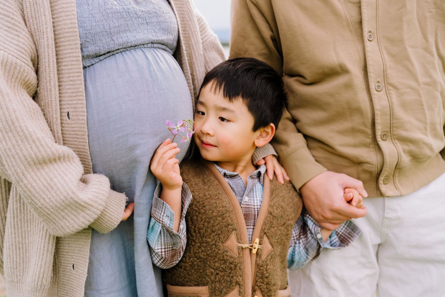 Young child in brown vest standing between two adults outdoors.