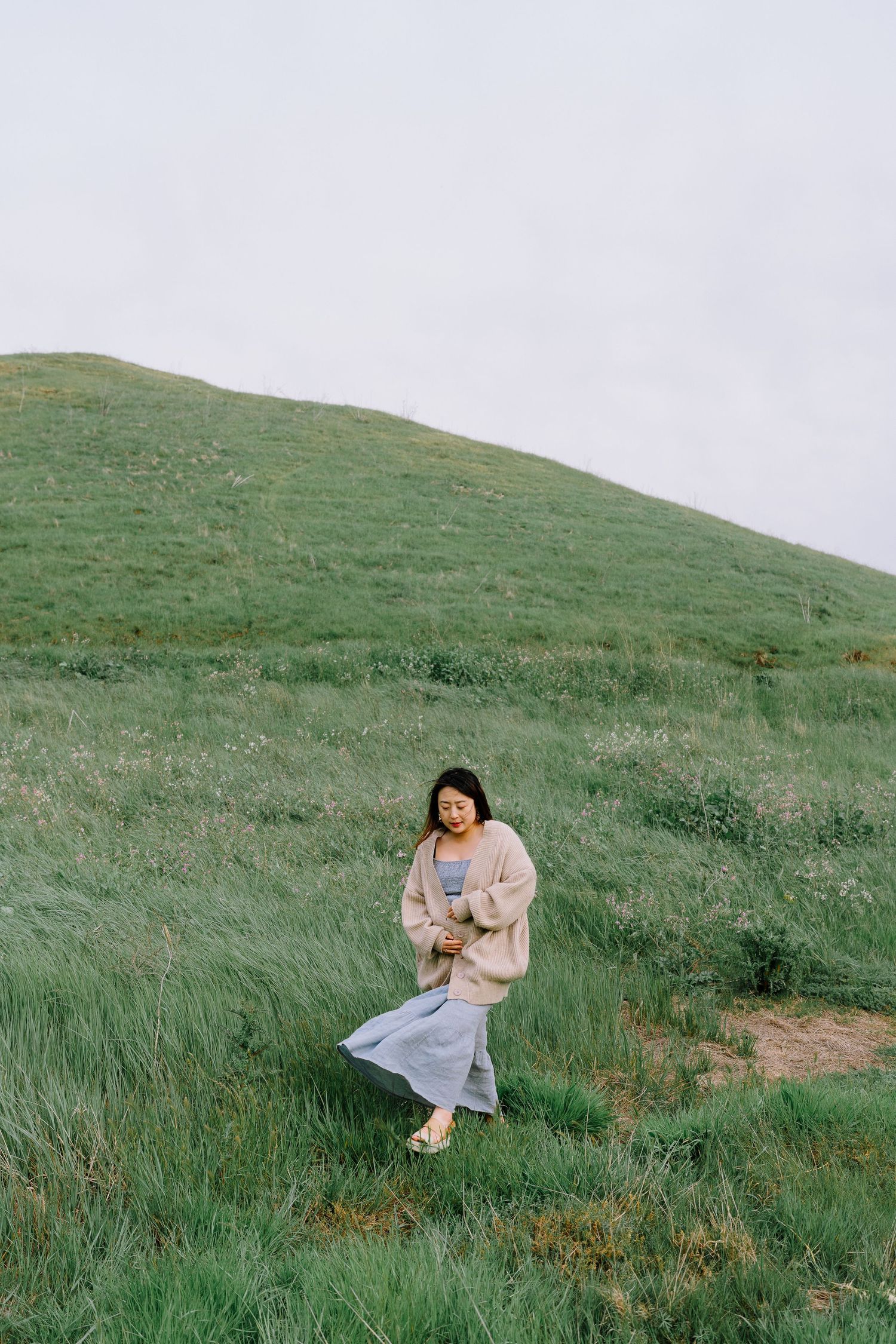 Person in casual attire sits peacefully on a grassy hillside against an overcast sky.
