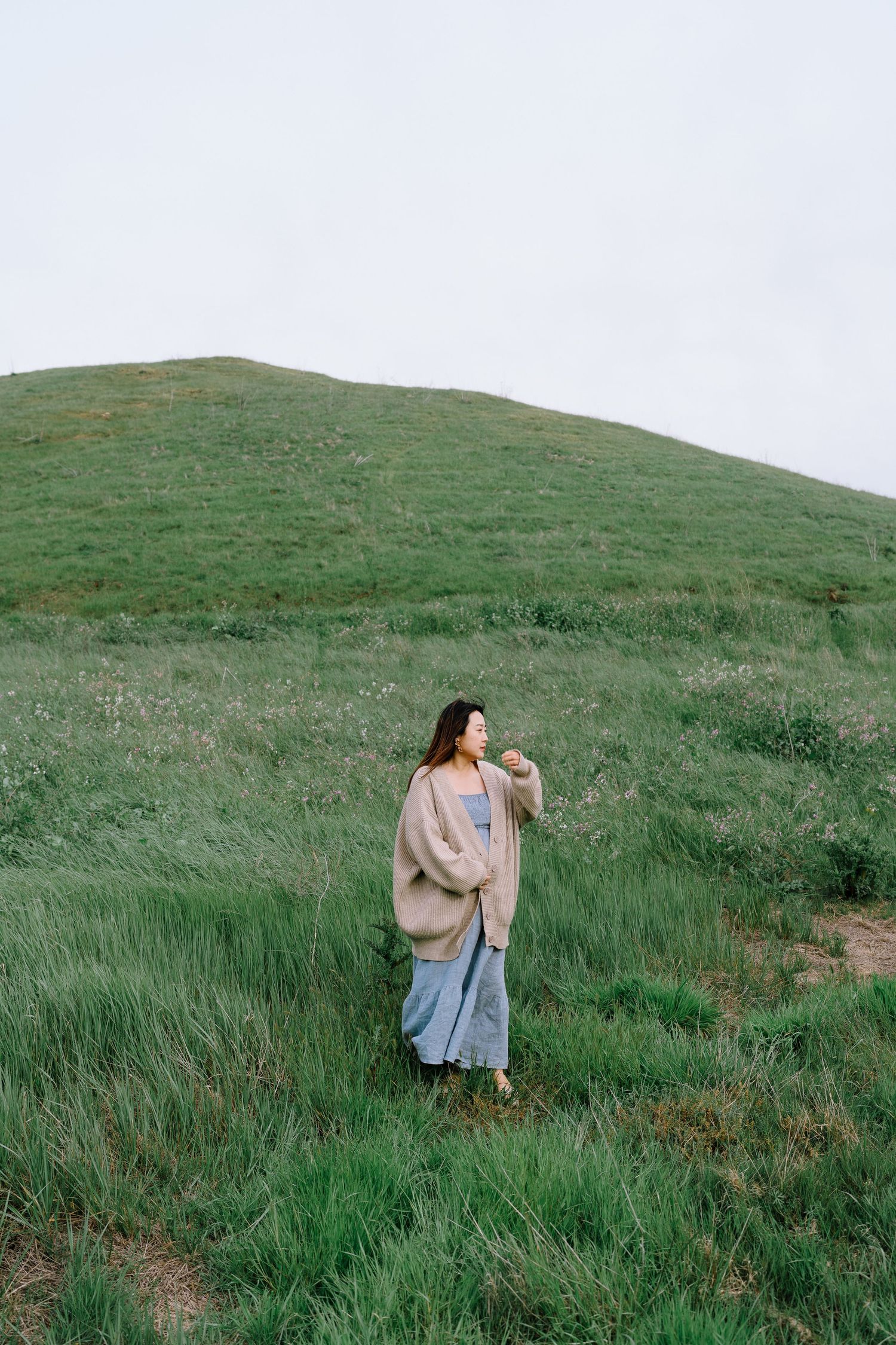 Person in oversized sweater stands on rolling green hillside covered in tall grass on overcast day.