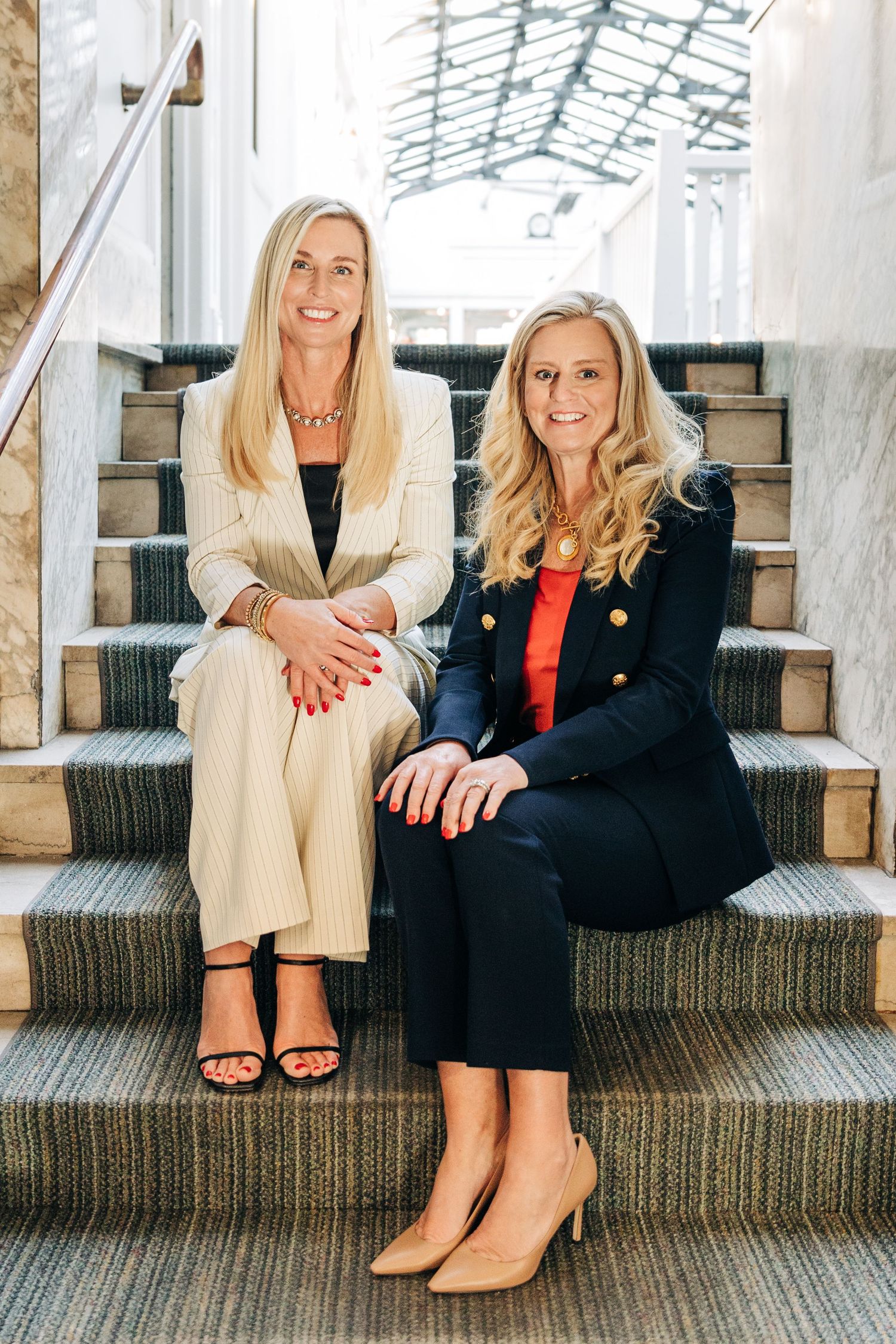 Two people in contrasting outfits sit on carpeted stairs in a bright indoor space with glass ceiling and stone pillars.