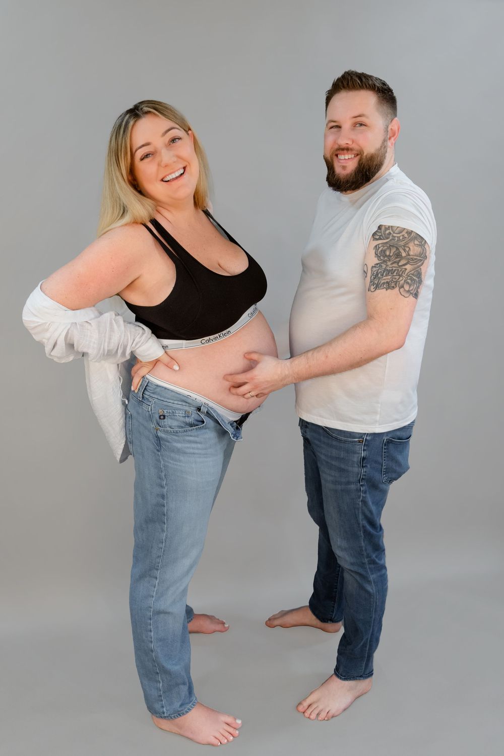 A playful couple in casual outfits share joyful moments during a photo session against a gray studio backdrop.