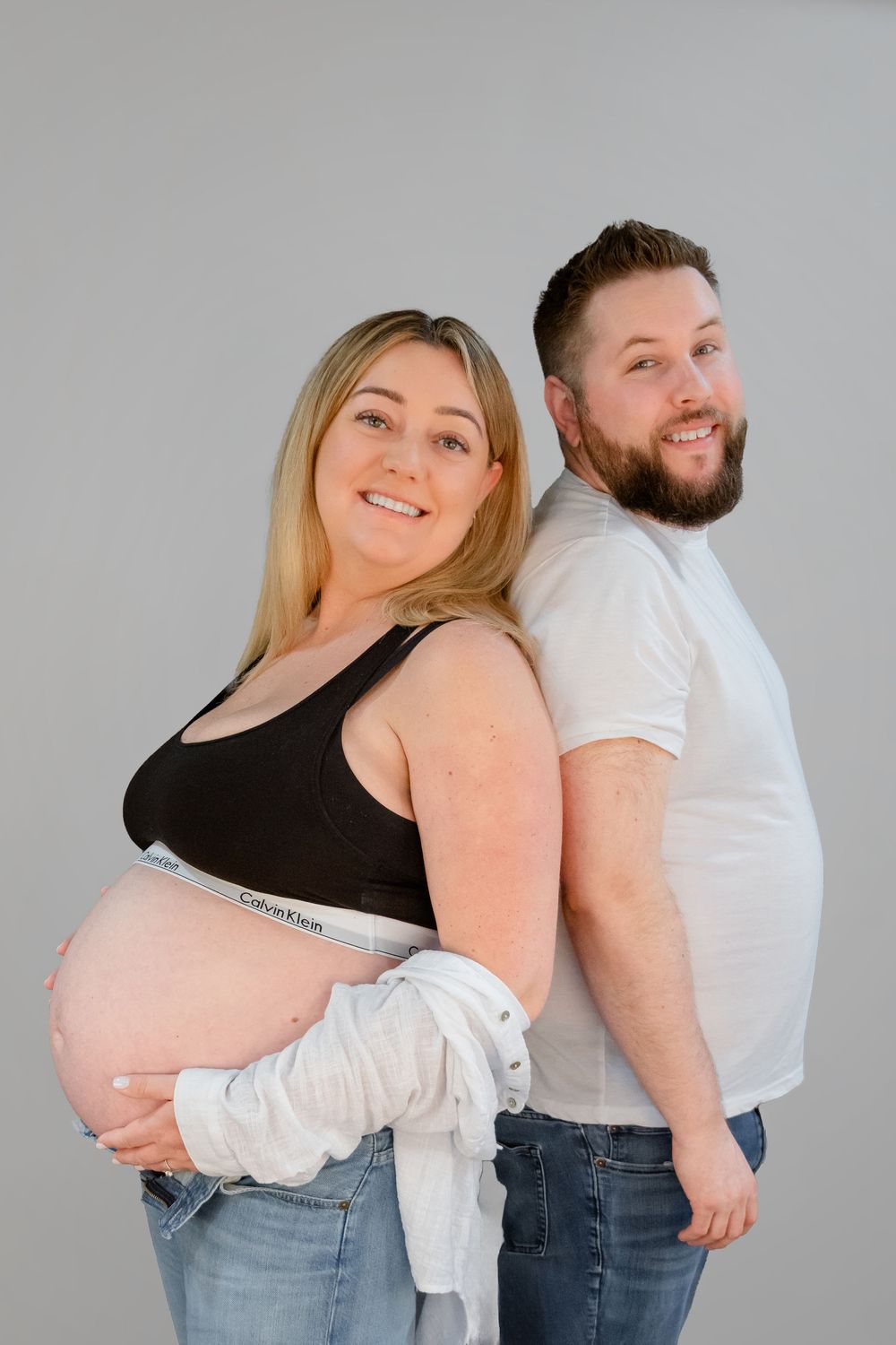 A couple poses back-to-back for a maternity photo against a gray background, wearing casual clothing.
