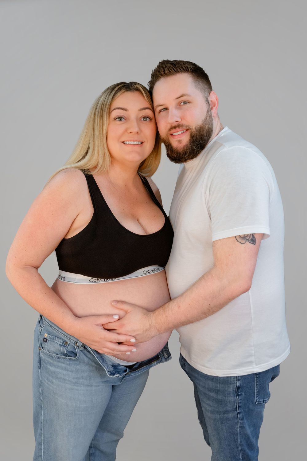 A couple in casual attire poses for a maternity photo against a gray background.