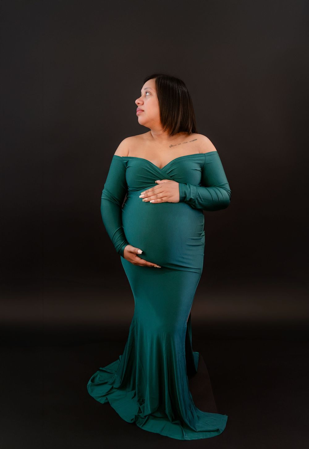 Artistic maternity photoshoot featuring an emerald green off-shoulder dress against a dark background.