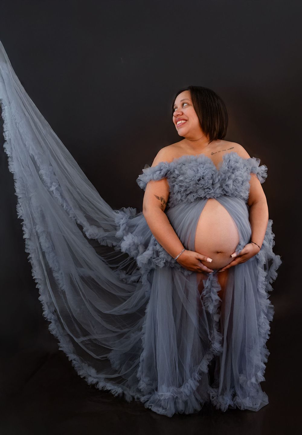 Maternity portrait in dusty blue tulle gown with flowing train against dark background.