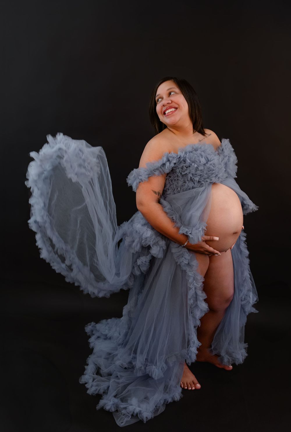 Maternity photos with flowing grey tulle skirt creating dramatic movement against black backdrop.