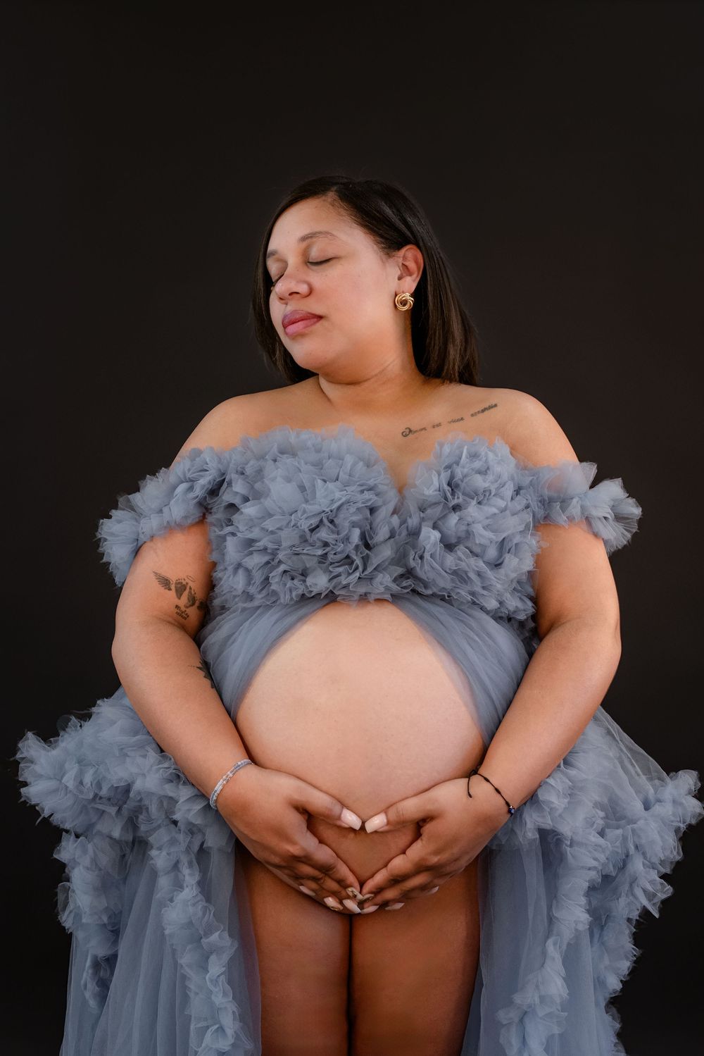 A maternity photo series showing a pregnant person in a flowing gray tulle gown against a dark background.