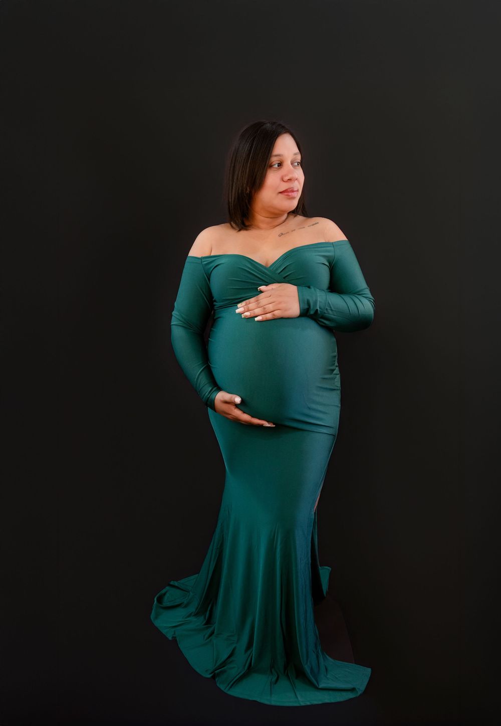 Woman in emerald green off-shoulder dress poses gracefully against dark background in maternity photoshoot series.