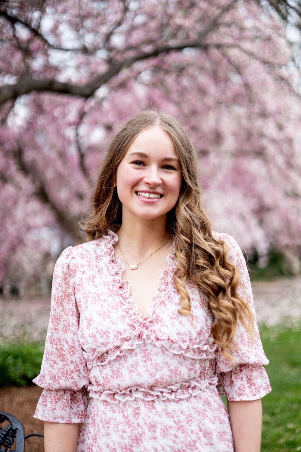 Woman in pink floral dress poses under blooming cherry blossom trees.