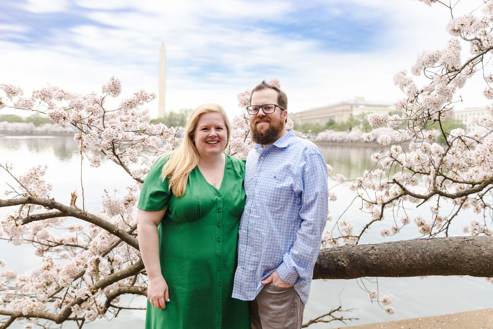 Couple poses together under cherry blossom trees near water on sunny spring day.
