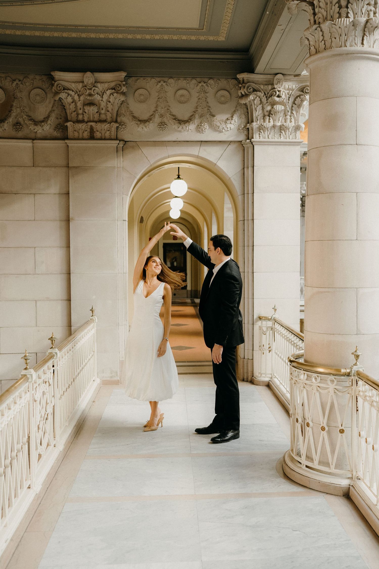 A couple in formal attire stands in an elegant arched hallway with ornate architectural details and classical columns.