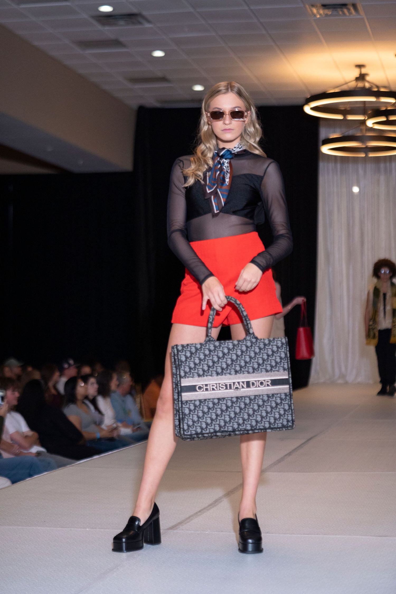 Fashion model walks runway in black mesh top, red skirt and platform shoes at fashion show event.