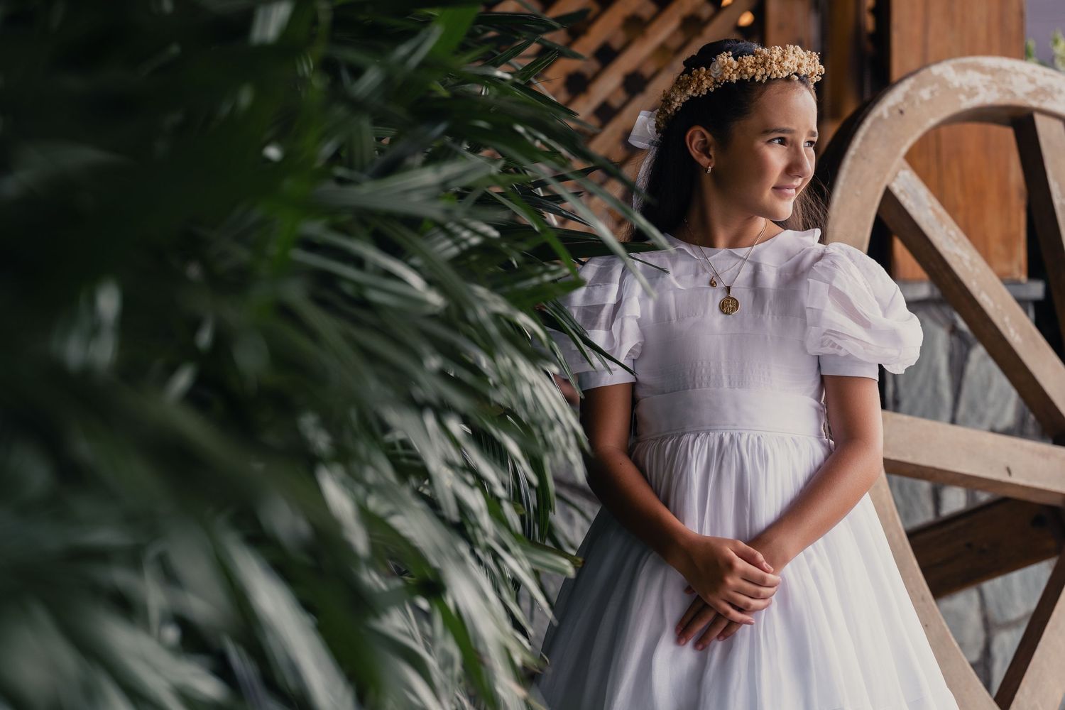 A young girl in a white communion dress with puff sleeves and tiara stands near tropical foliage and wooden architecture.
