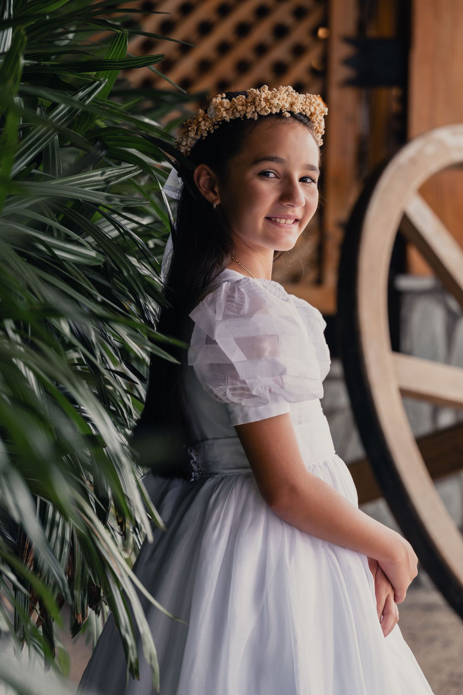 A young child in a white formal dress with ruffled collar poses near tropical plants at an elegant venue.