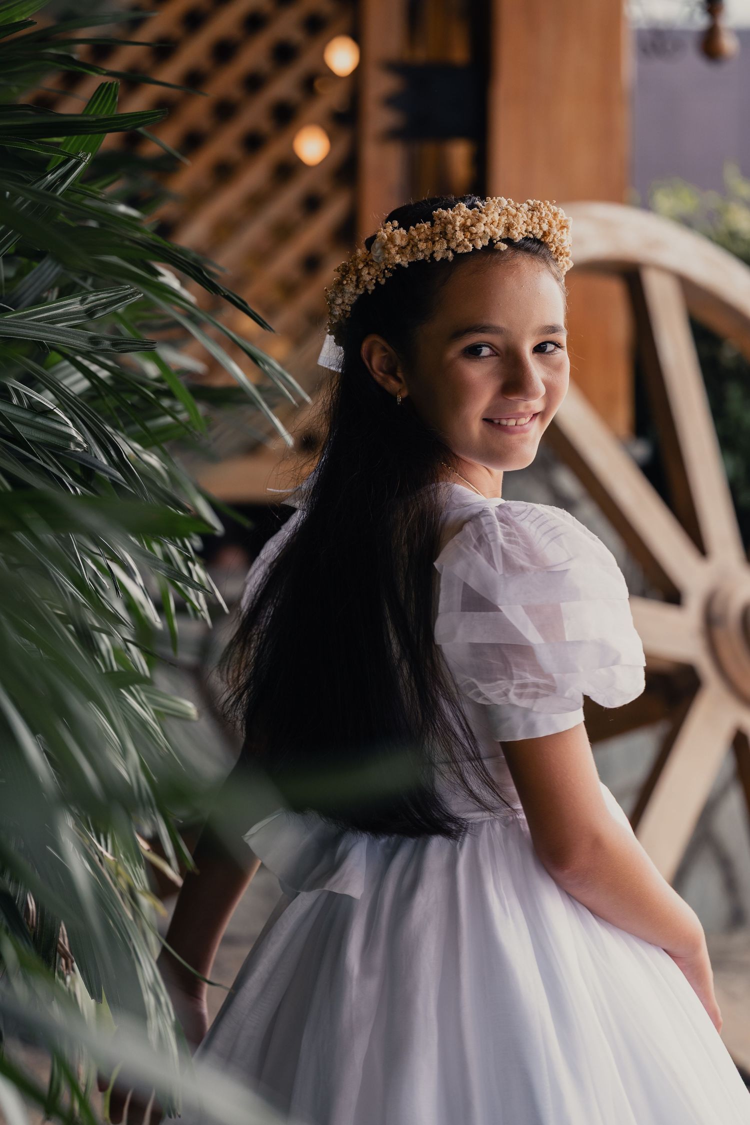 Young person in white communion or special occasion dress with floral crown and long dark hair poses by tropical plants.