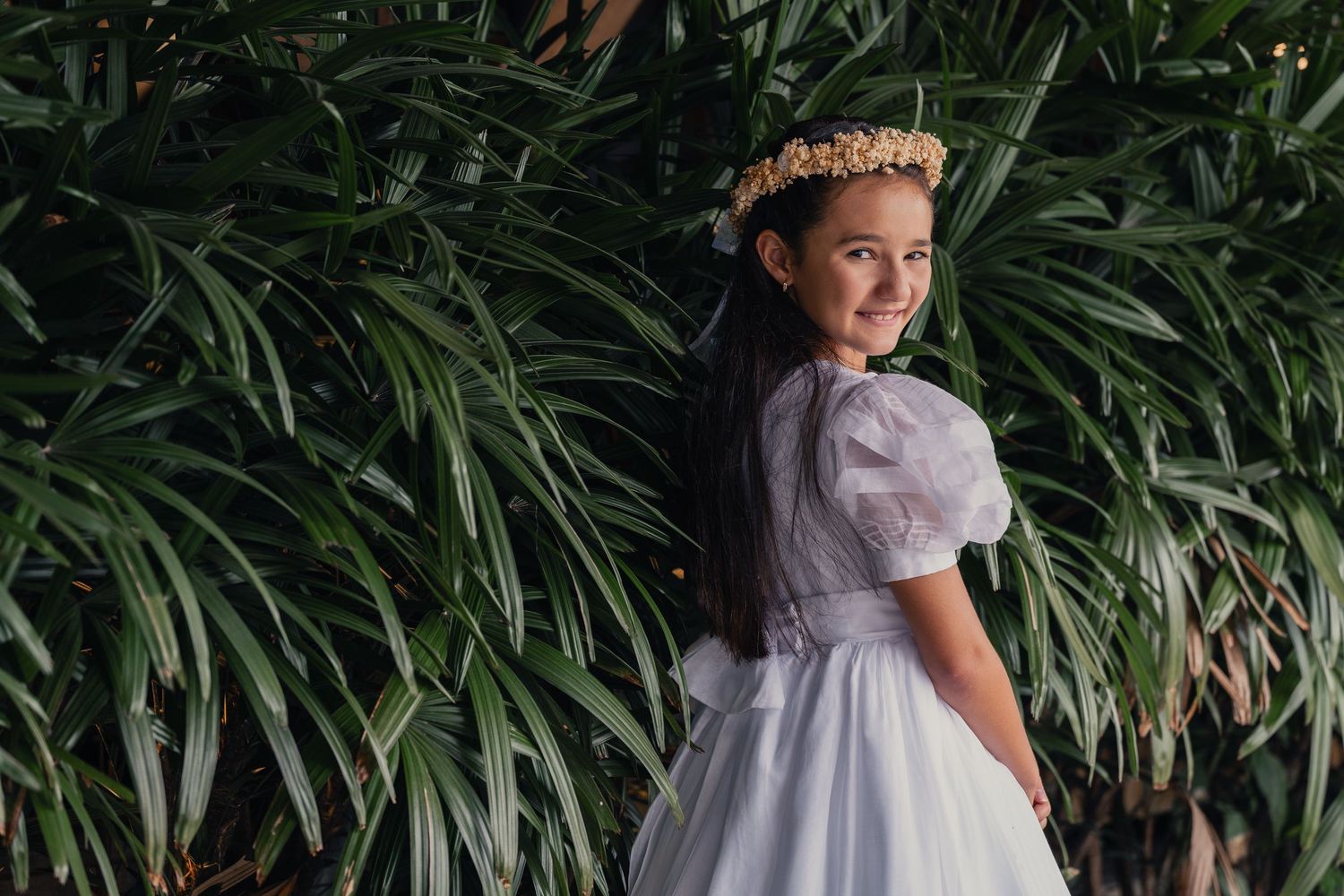 A person in a white dress stands among lush tropical greenery in an outdoor garden setting.