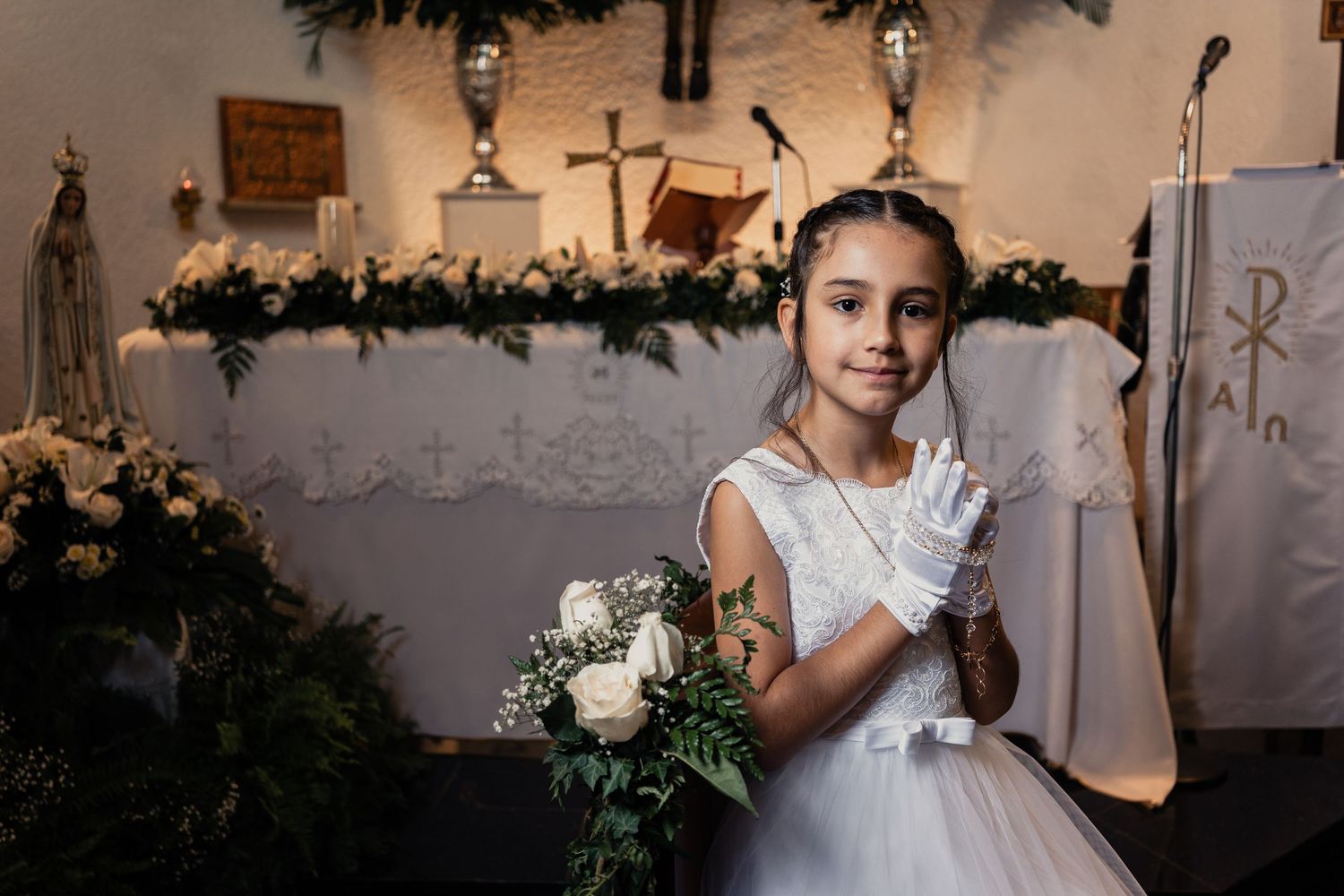 A young person in a white First Communion dress holds a bouquet and candle in a church decorated with floral garlands.